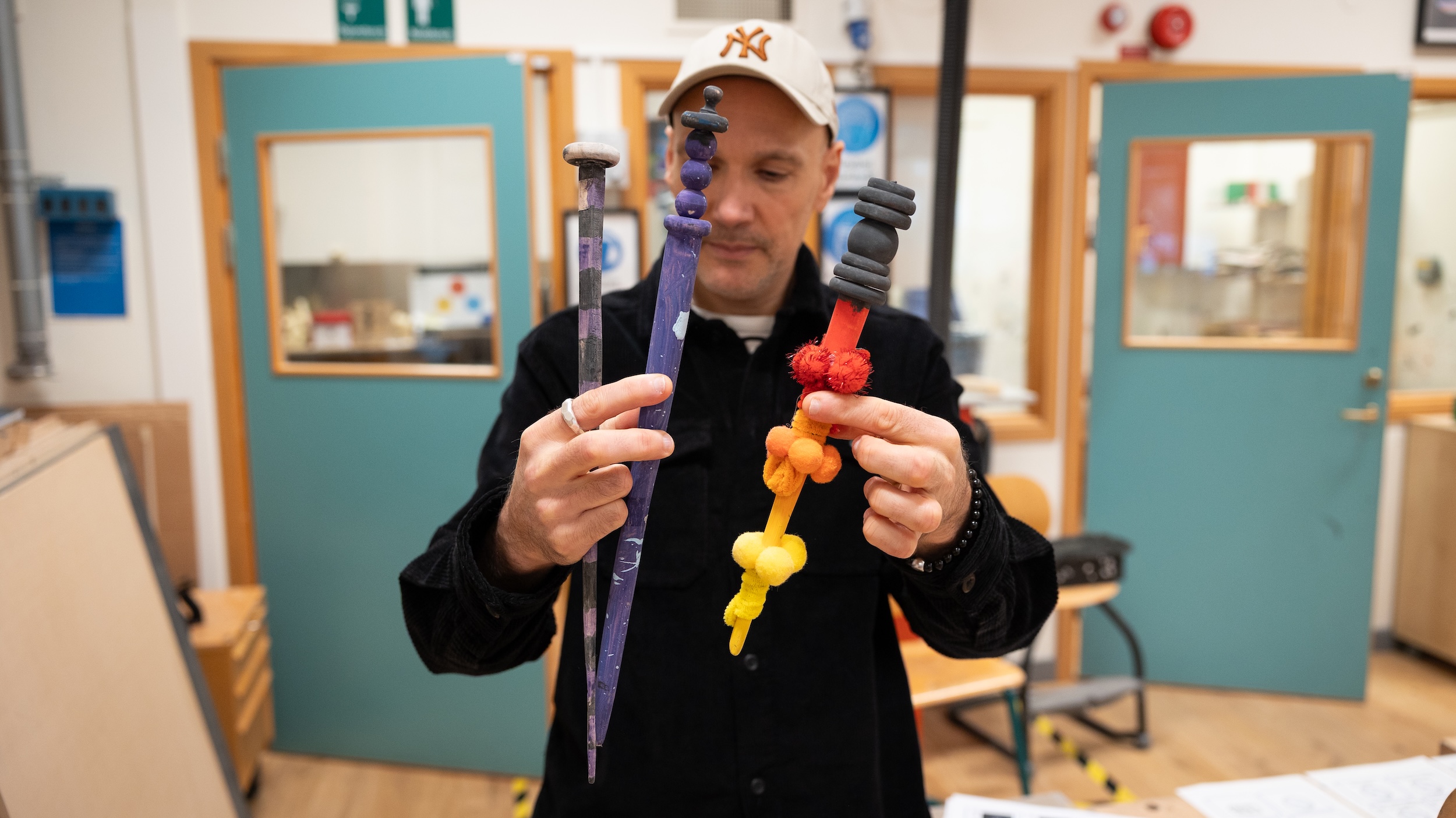 Person i slöjdsal håller i två färgglada trollstavar som fjärdeklassare har tillverkat. Bakgrunden visar dörrar och verktyg i en skolmiljö för träslöjd.