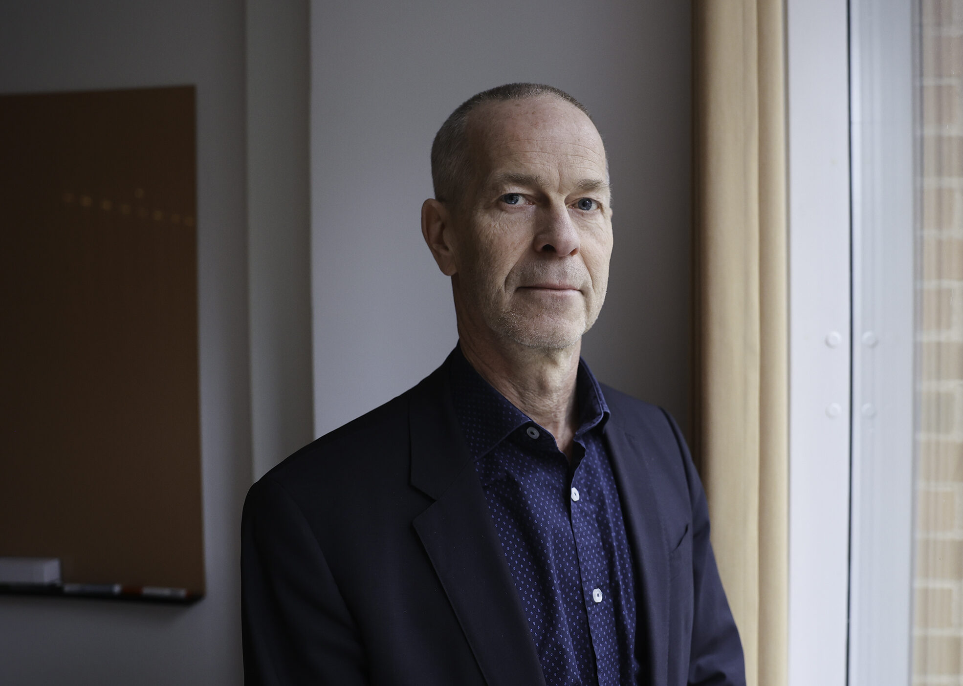 Man i kavaj står nära fönster och tittar in i kameran.