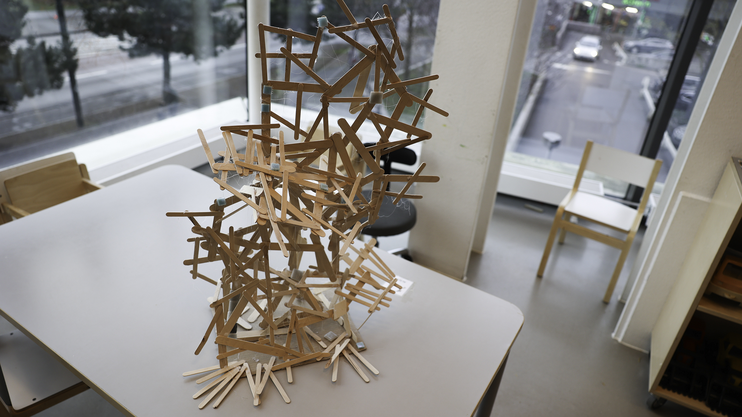 Stor konstruktion byggd av tunna träpinnar står på ett bord i ett klassrum med stora fönster mot en gata.