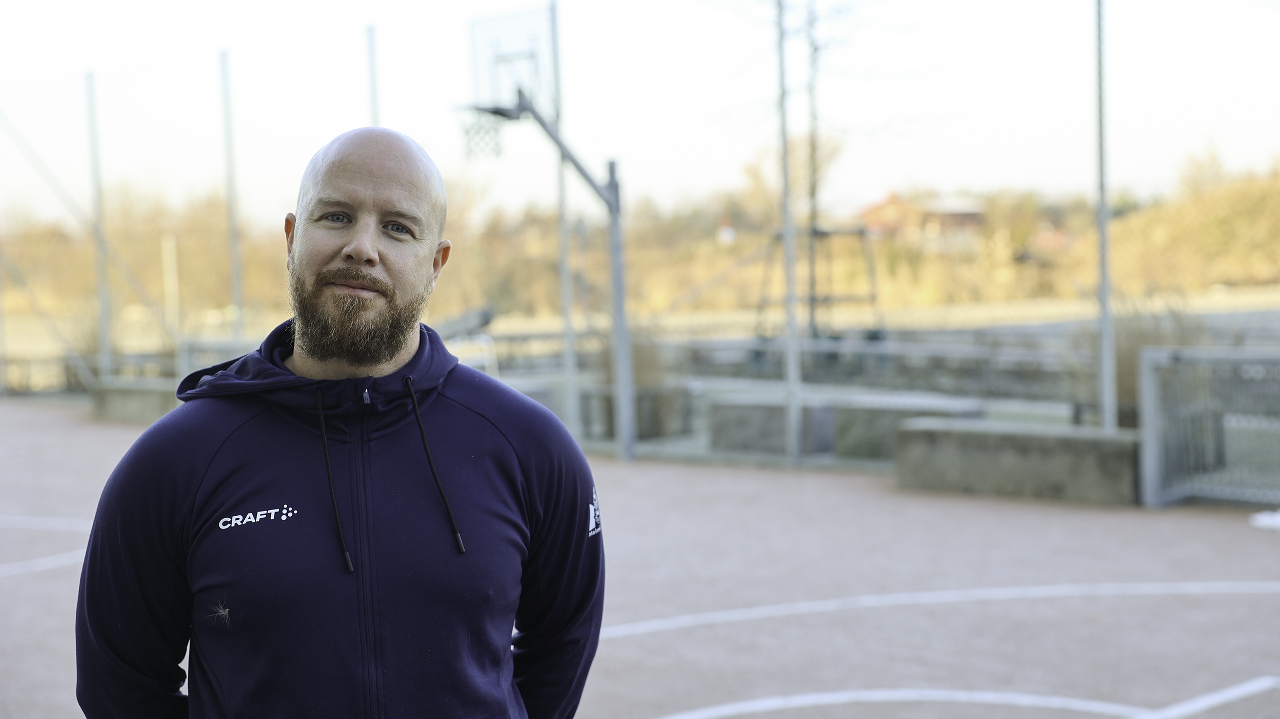Person i sporttröja står på en utomhusplan med basketkorgar och stängsel i bakgrunden.