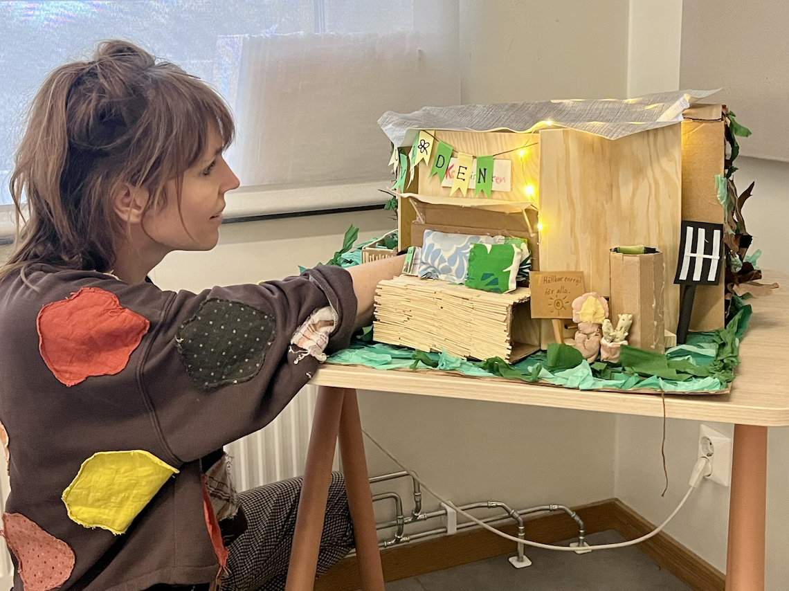 Lärare arbetar tillsammans med en elevbyggd modell av en hållbar miniatyrmiljö, dekorerad med naturmaterial och ljusslingor, som en del av ett kreativt och ämnesintegrerat projekt i skolan.