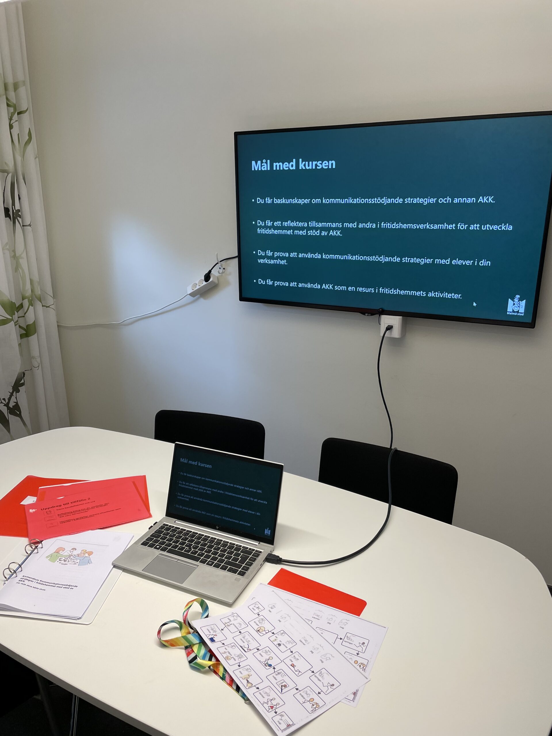 Mötesrum med laptop, kursmaterial och färgmarkerade pärmar på ett bord, medan en skärm på väggen visar målen för en kurs om kommunikationsstöd och AKK.