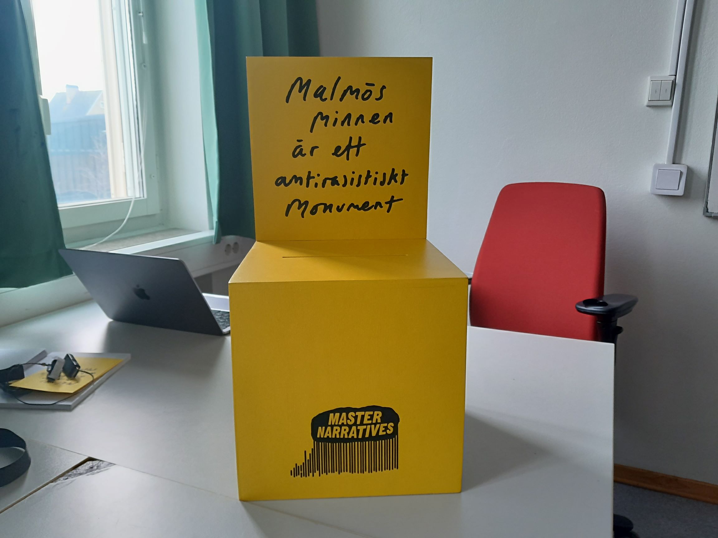 Gul kub med texten “Malmös minnen är ett antirasistiskt monument” och logotypen Master Narratives stående på ett skrivbord i ett kontorsrum med laptop och röd stol i bakgrunden.