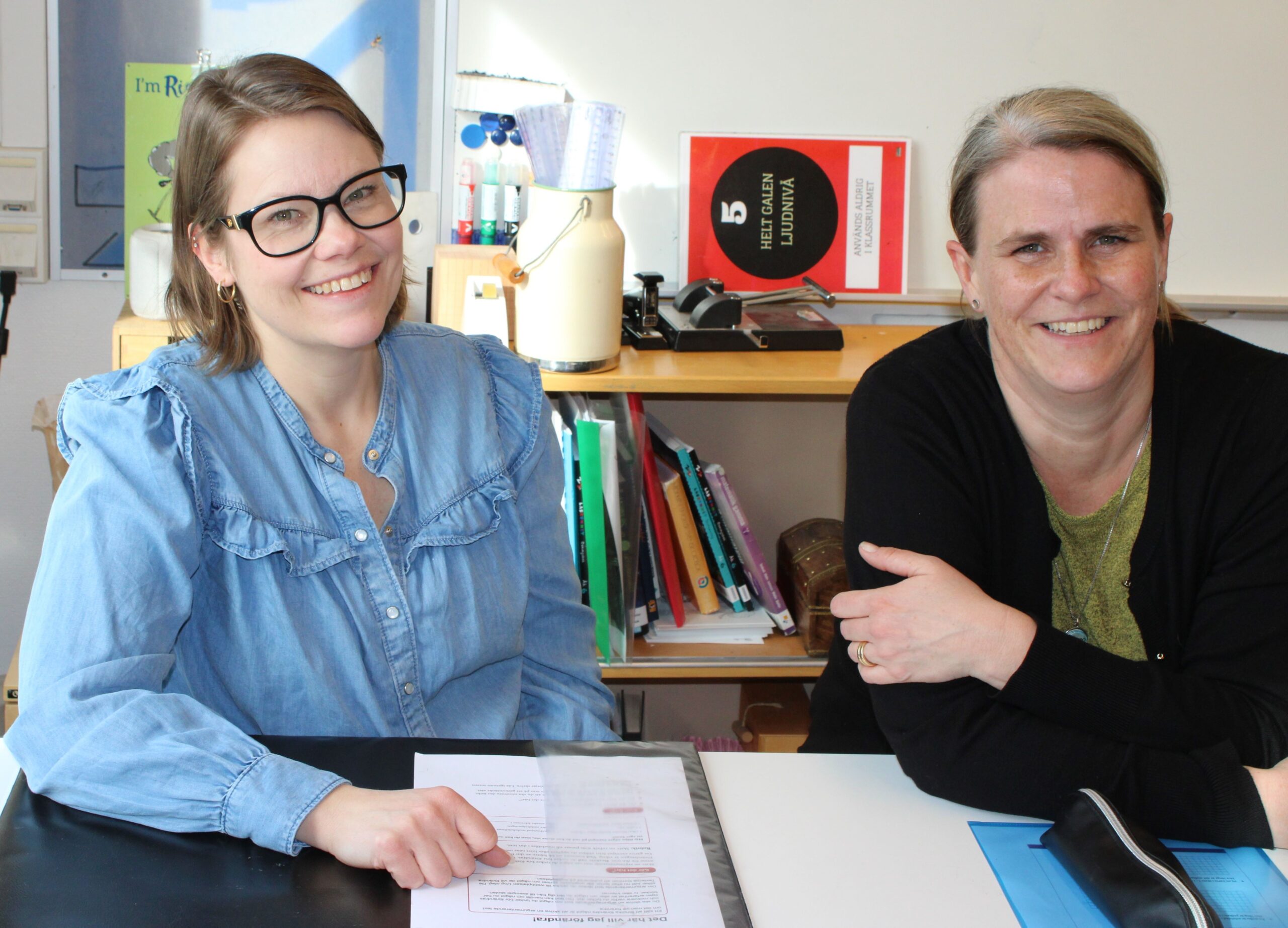 Två personer sitter vid ett bord med papper och mappar i ett rum med hylla och material i bakgrunden.