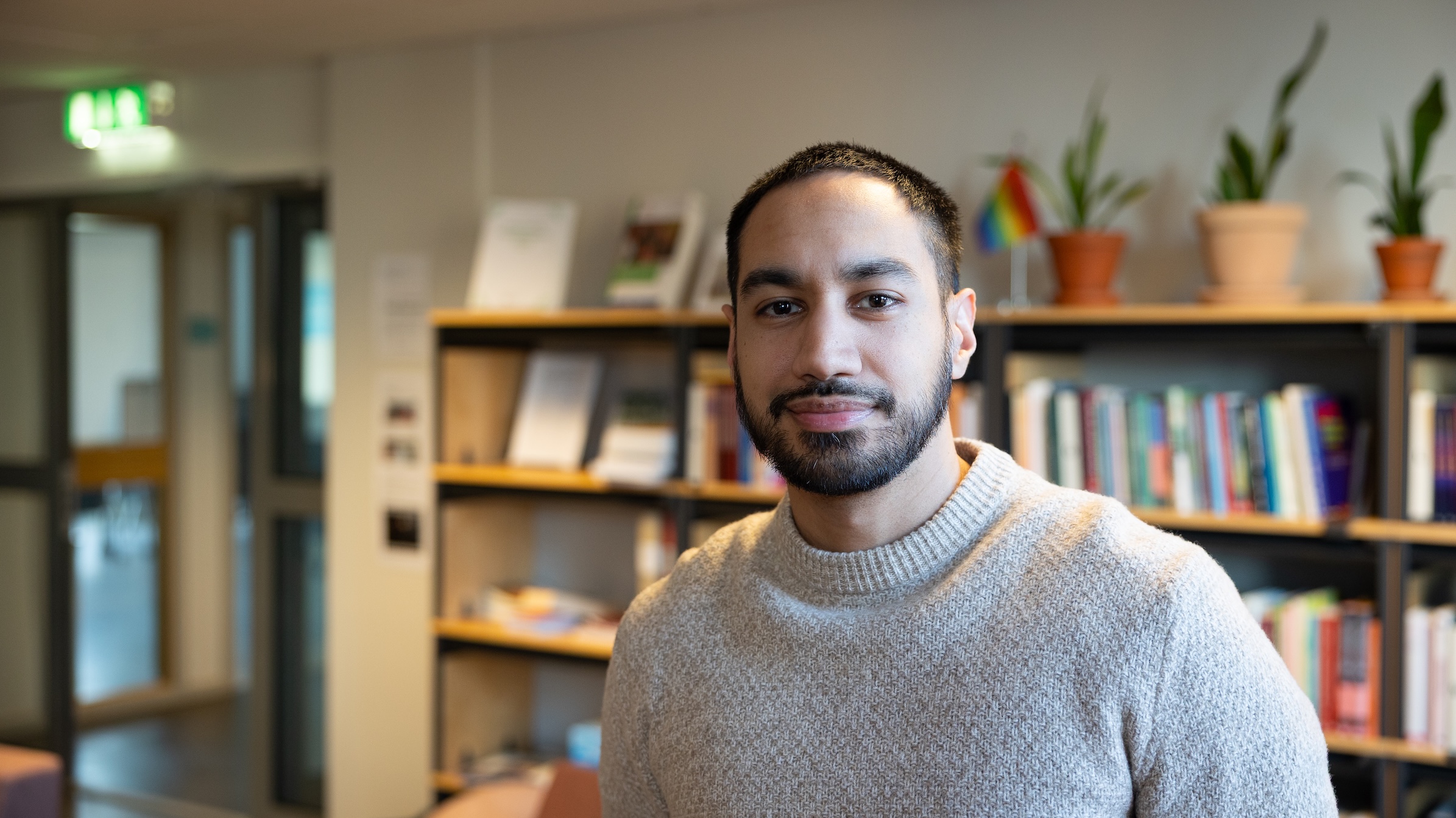 Person i ljus tröja i ett rum med bokhyllor och växter i bakgrunden.