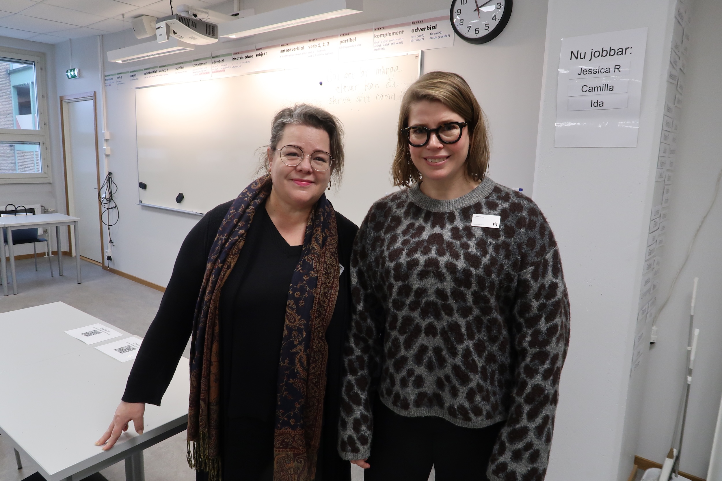 Två personer står i ett klassrum framför en whiteboard med schema och instruktioner. I bakgrunden syns bord, stolar, en väggklocka och en skylt med texten ”Nu jobbar: Jessica R, Camilla, Ida”.