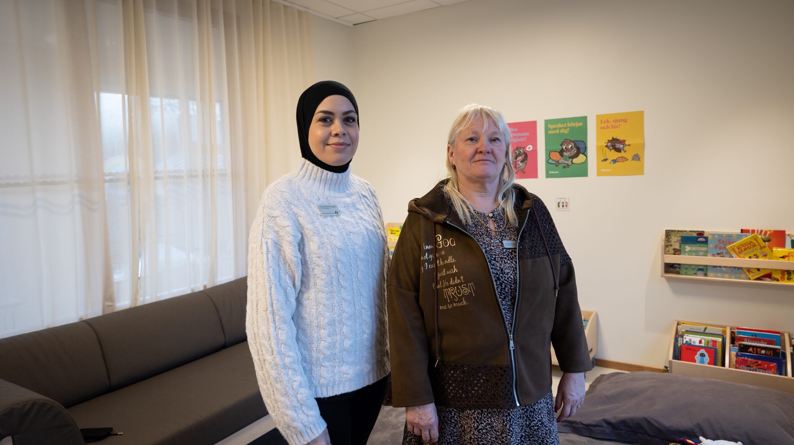Två pedagoger står i ett ljust förskolerum med soffa, bokhylla och färgglada affischer på väggen. De bär namnskyltar och står bredvid varandra i miljön där elevernas böcker och material är synligt organiserade.