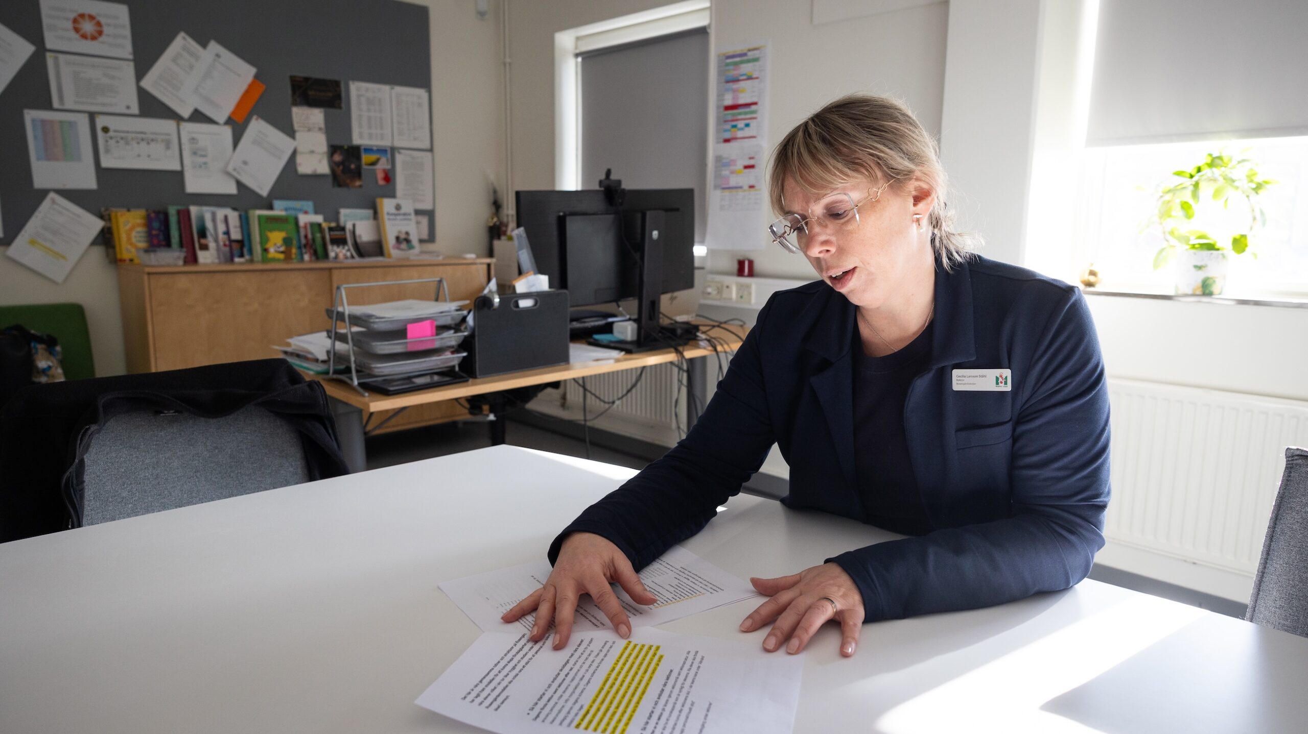 Person sitter vid ett bord i ett kontorsliknande arbetsrum och går igenom utskrivna dokument med markerad text. Bakgrunden visar hyllor, pärmar, en dator, anslagna papper på väggen och ett fönster med växt på fönsterbrädet.