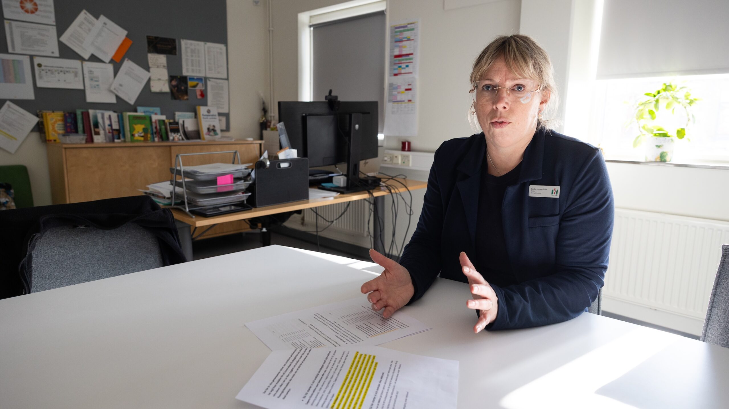 Person sitter vid ett bord i ett ljust arbetsrum och förklarar innehållet i utskrivna dokument med markerad text. Bakom personen syns en datorarbetsplats, hyllor med böcker och papper samt en anslagstavla med uppsatta dokument.