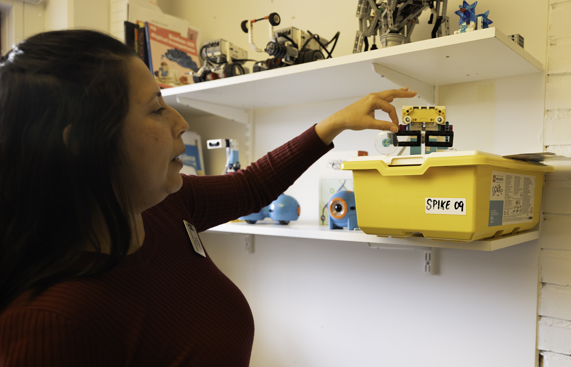 Person visar en liten robotmodell från en hylla i ett makerspace‑ eller teknikrum, där flera programmerbara robotar och en gul låda märkt “Spike 09” står uppställda.