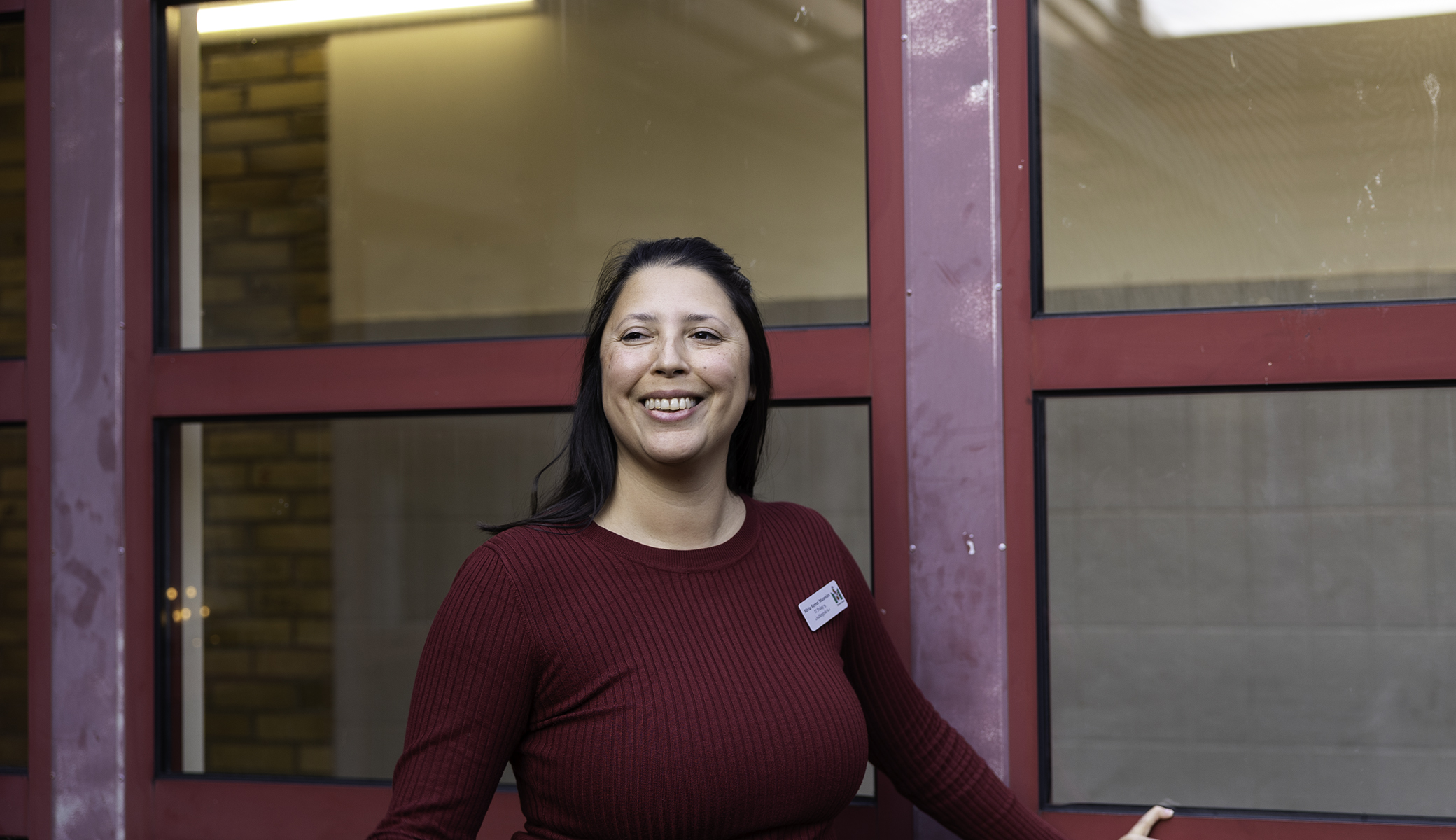 Person står framför en röd fönsterfasad inomhus och bär en vinröd tröja och namnskylt, i en skolmiljö med tegelväggar och glaspartier i bakgrunden.