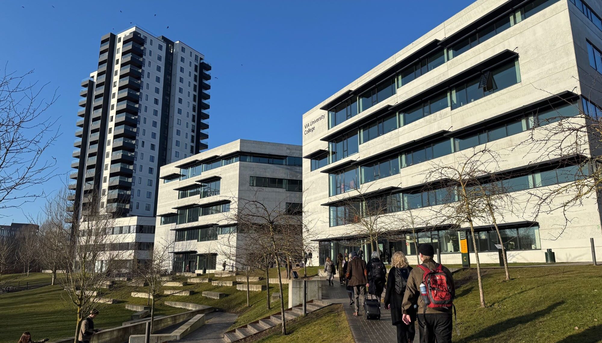 Personer promenerar på gångväg framför moderna fler­våningsbyggnader i soligt väder.