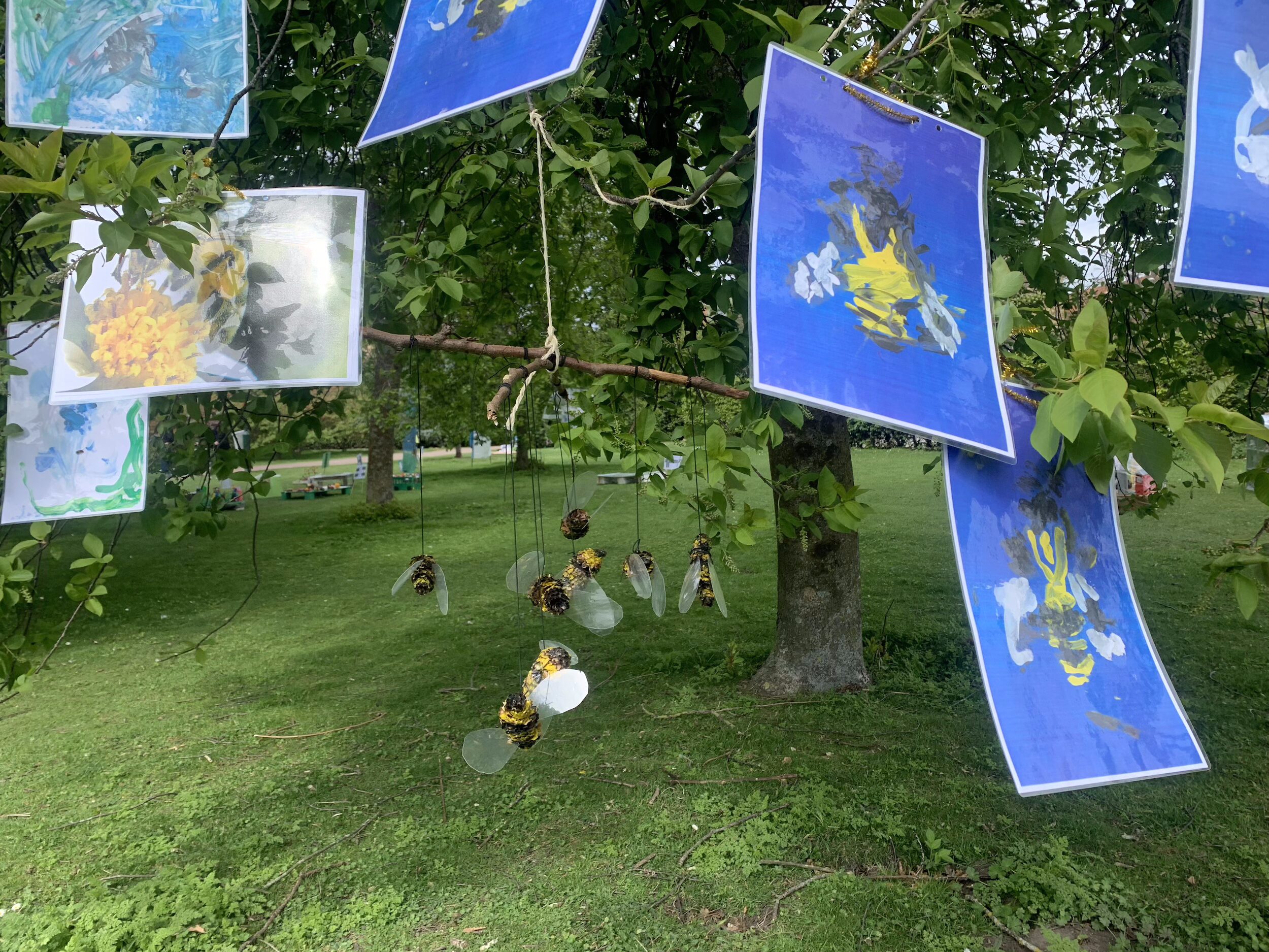 Utomhusinstallation med bilder och målningar upphängda i ett träd samt hängande figurer, placerad i en grön parkmiljö.