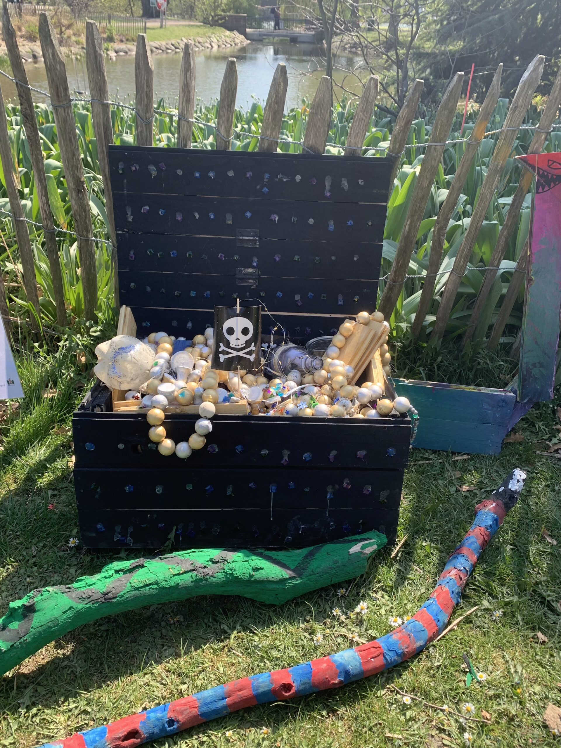 Öppen svart skattkista utomhus fylld med pärlor och föremål, dekorerad med dödskallesymbol och placerad vid ett staket nära vatten.