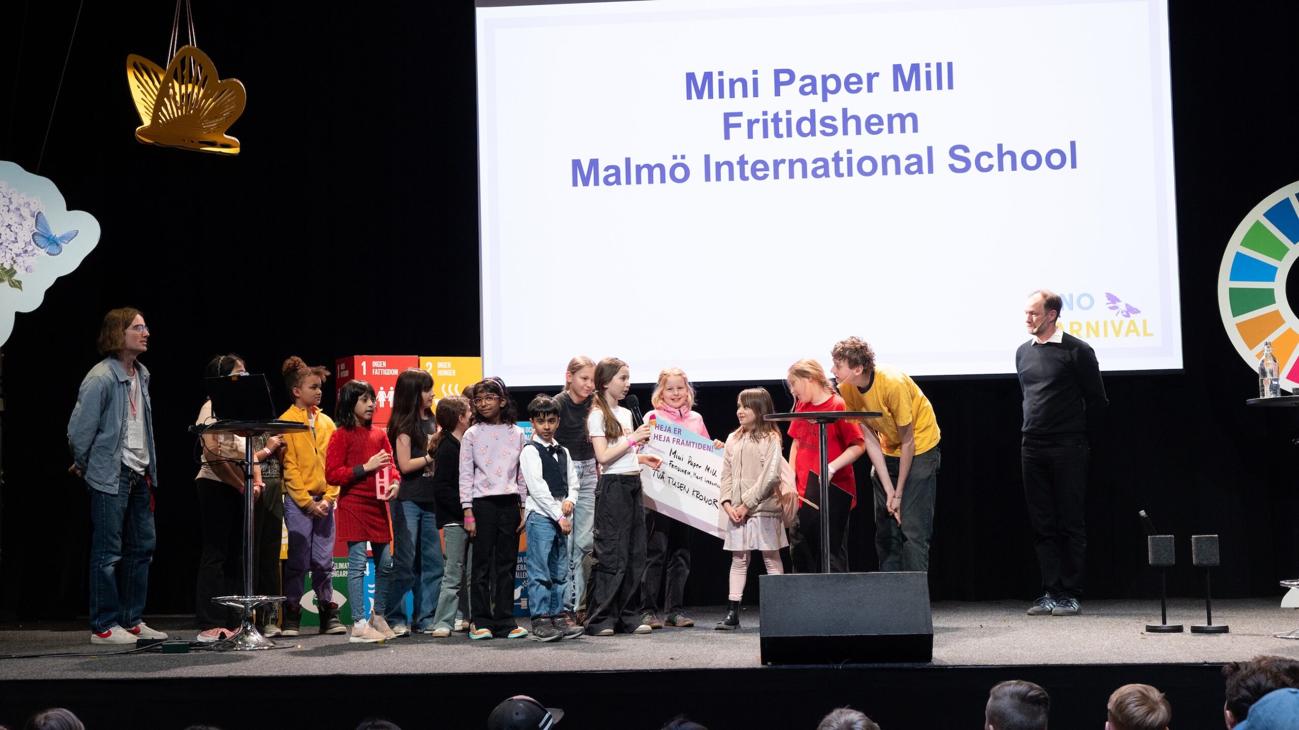 Grupp barn och vuxna står på en scen framför en stor skärm med texten Mini Paper Mill, Fritidshem, Malmö International School.