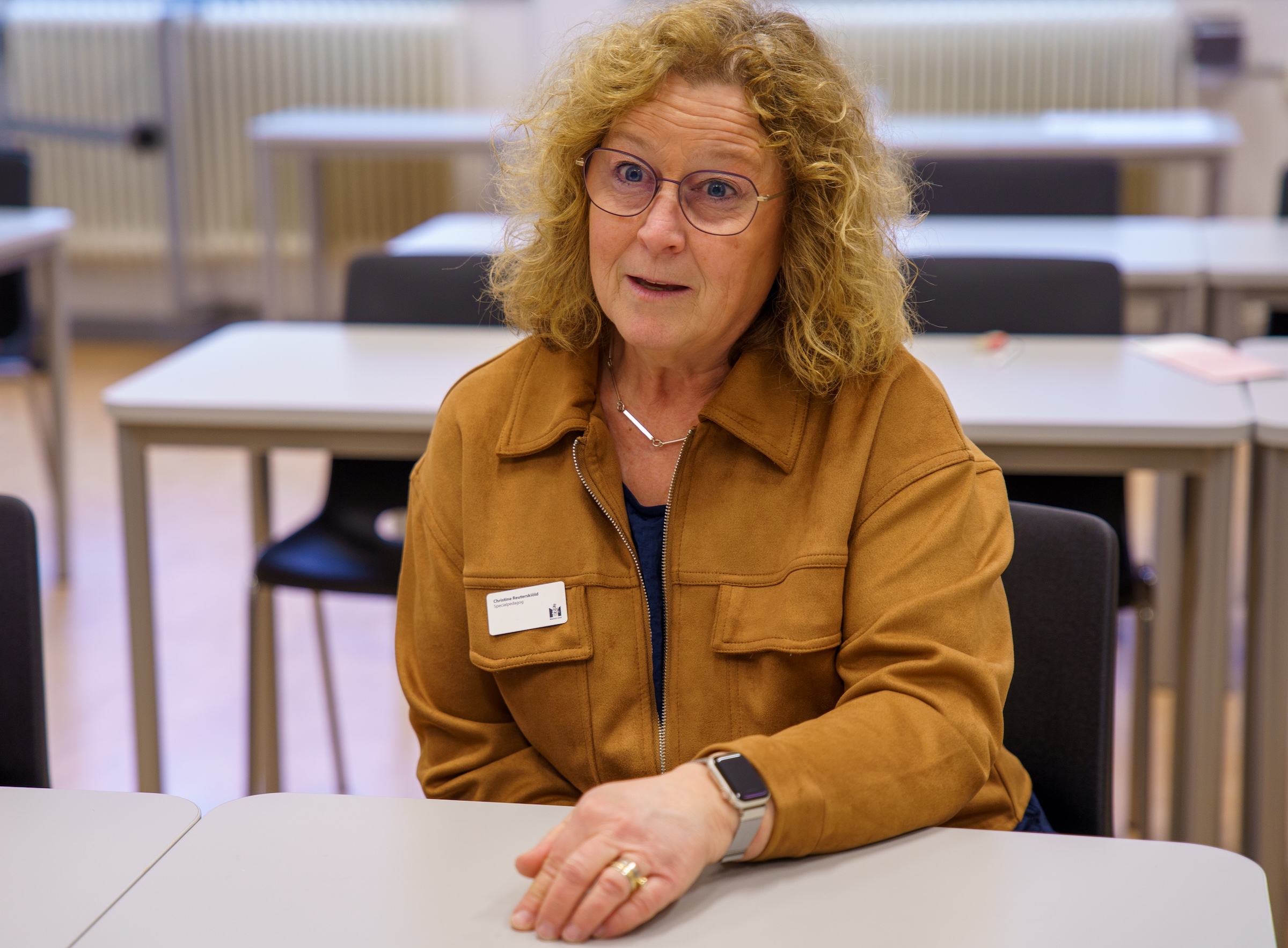 Person sitter vid ett bord i ett klassrumsmiljö med stolar och bord i bakgrunden.