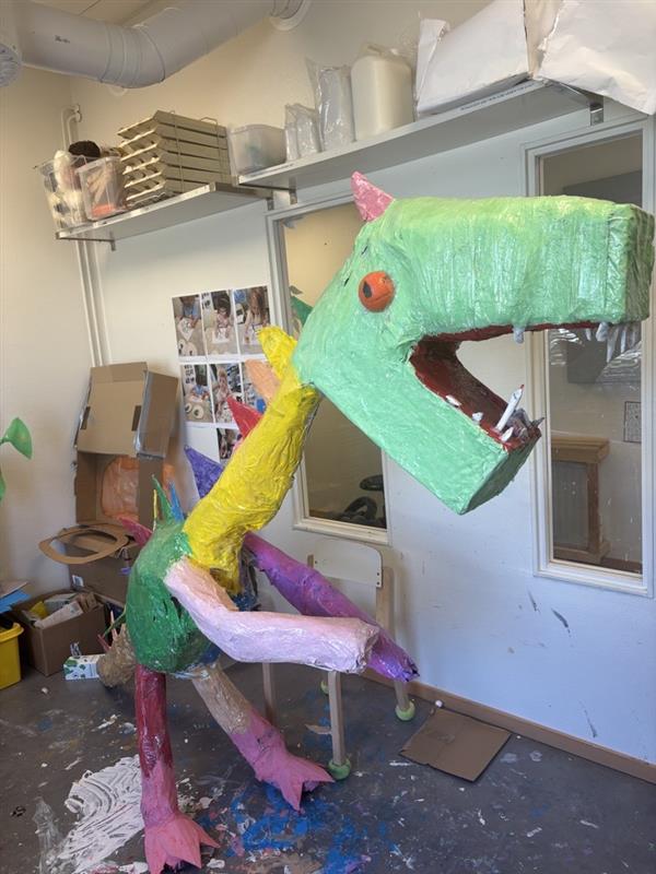 Färgstark handgjord skulptur av ett fantasidjur med stort öppet gap, placerad i ett skapanderum med material och hyllor i bakgrunden.