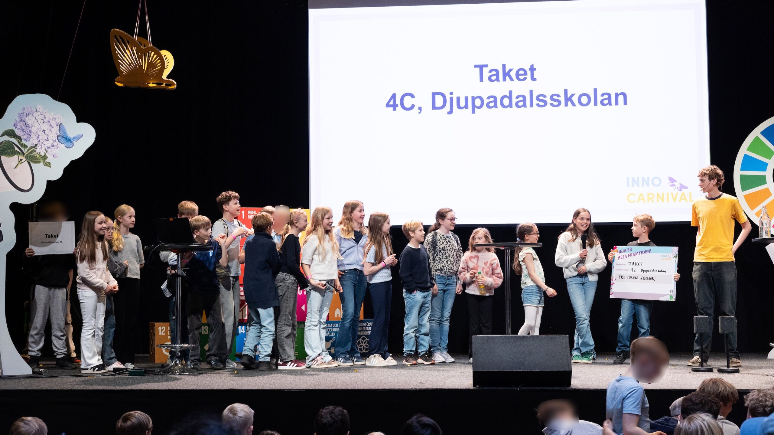 Grupp barn står på en scen framför en stor skärm med texten Taket, 4C, Djupadalsskolan, tillsammans med skyltar och scenutrustning.