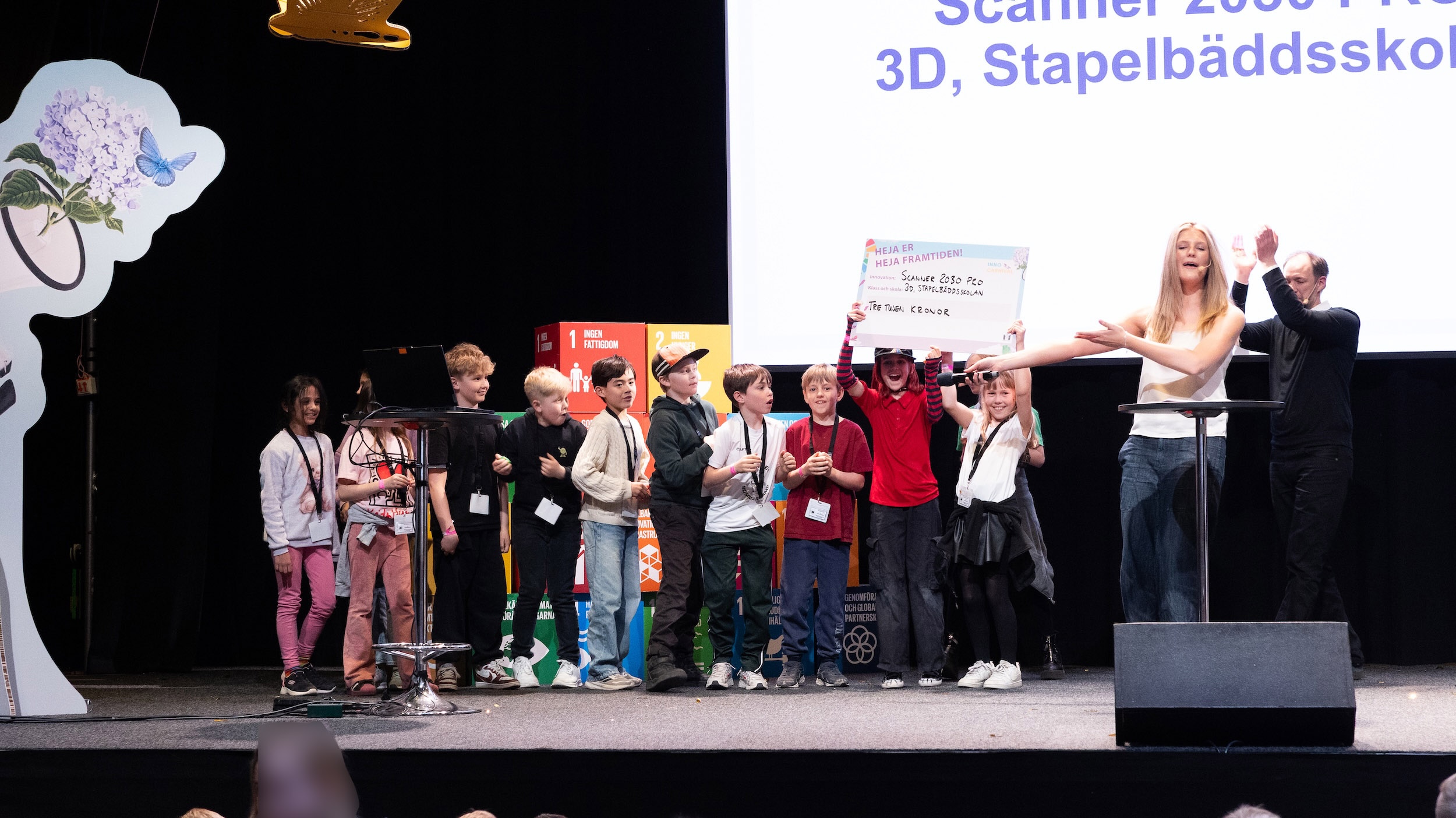 Grupp barn och vuxna står på en scen och håller upp en skylt framför en stor presentationsskärm med text om 3D och Stapelbäddsskolan.