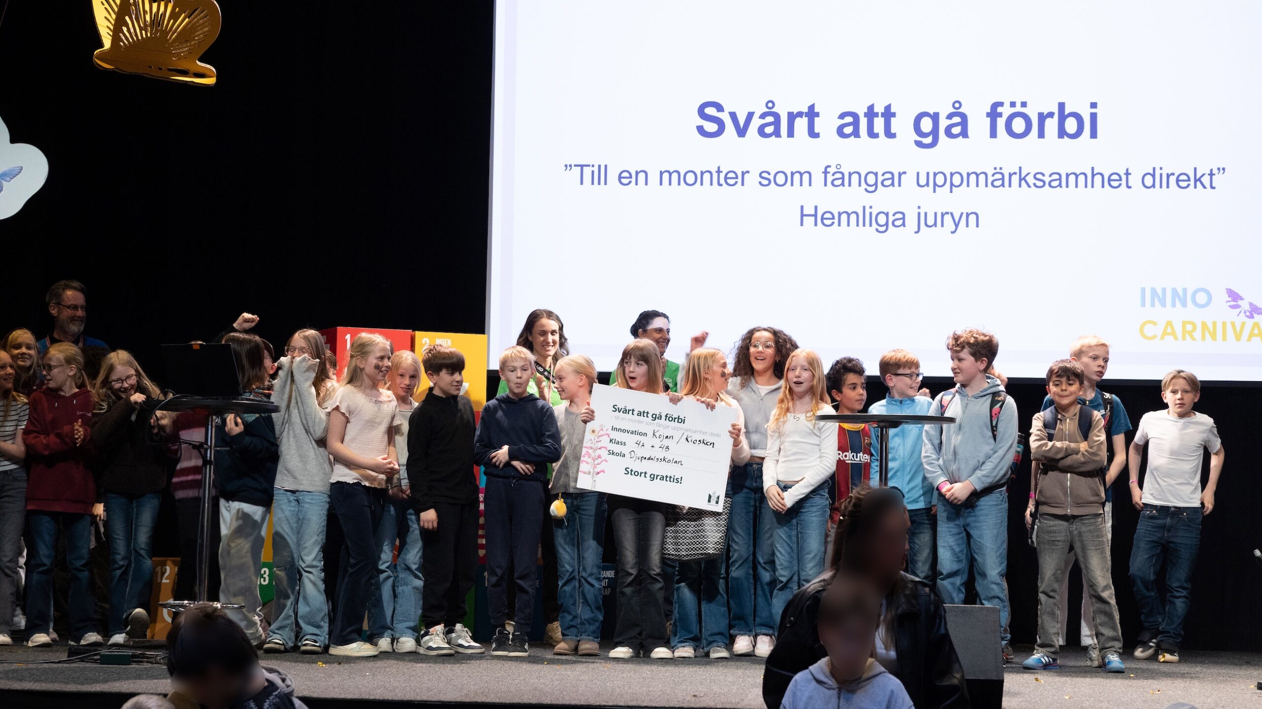 Grupp barn står på en scen framför en stor skärm med texten Svårt att gå förbi och InnoCarnival, där en skylt hålls upp.