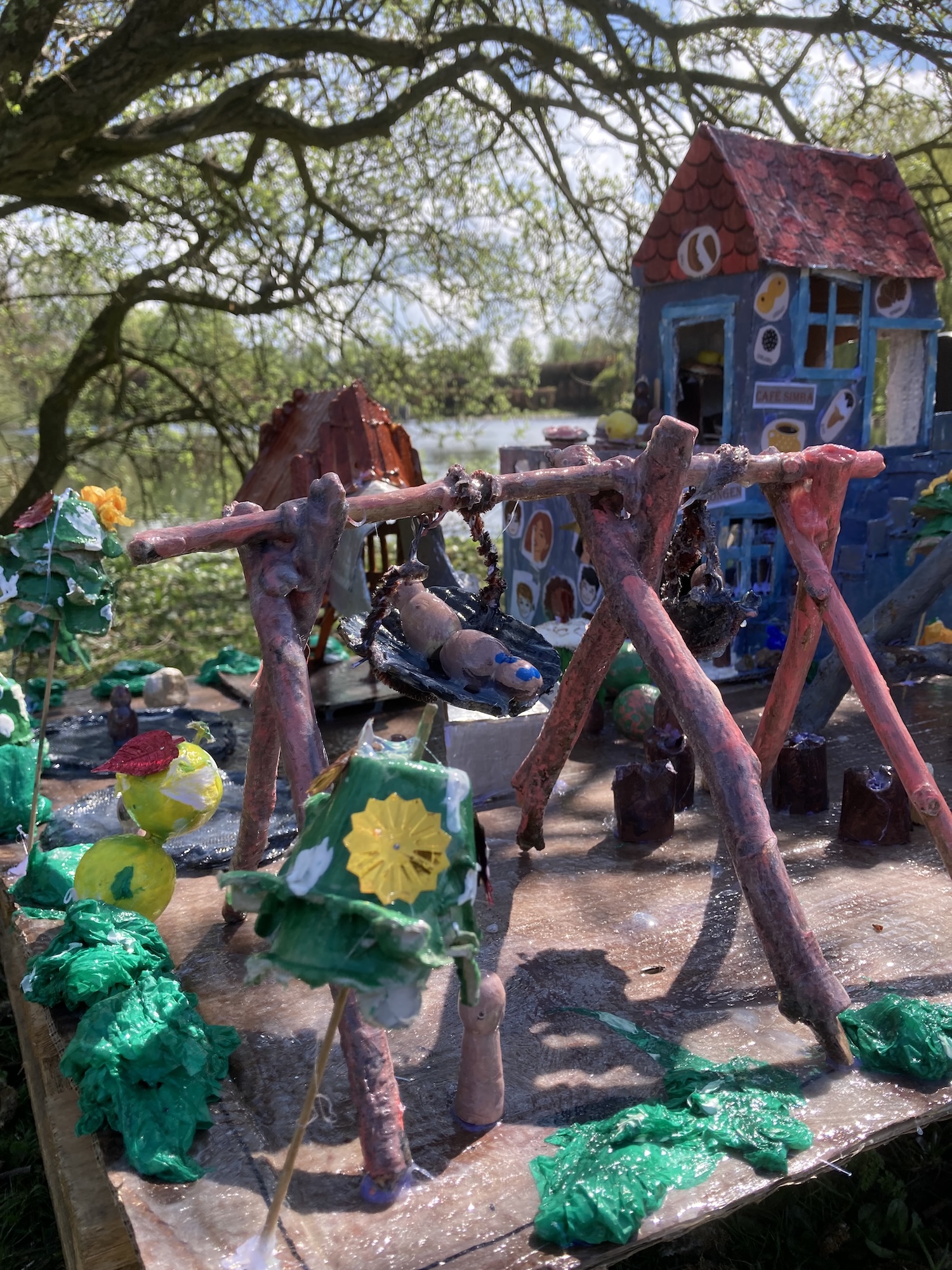 Handgjord miniatyrmiljö utomhus med små hus, figurer och konstruktioner av naturmaterial placerade vid ett vattendrag.