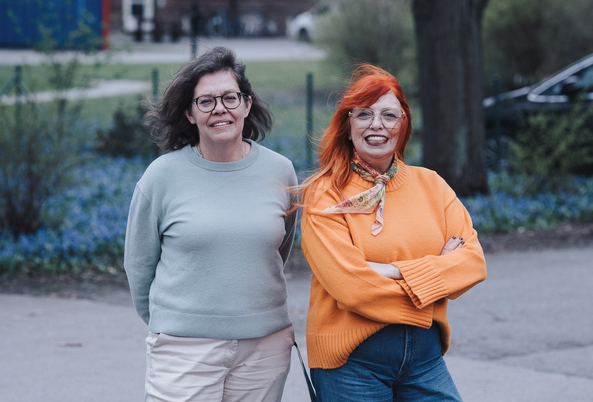 Två personer står utomhus på en gångväg med parkmiljö, träd och blomsterplanteringar i bakgrunden.