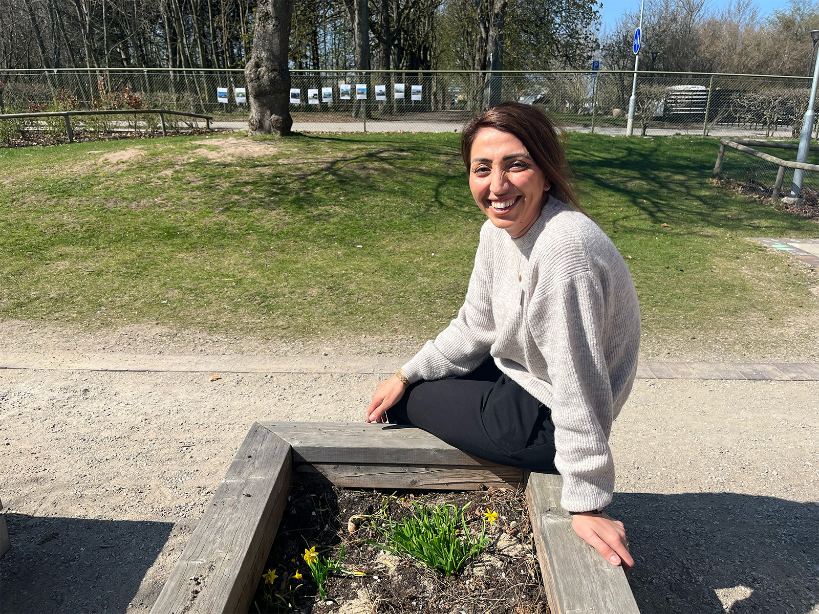 Person sitter utomhus vid en upphöjd planteringslåda i en parkmiljö med gräsytor och träd i bakgrunden.