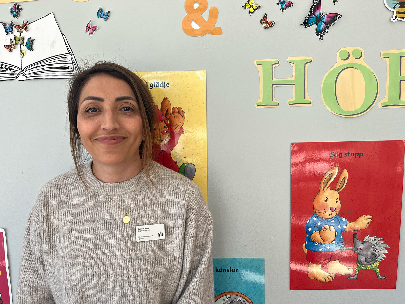 Person står inomhus framför en vägg med barnboksaffischer, fjärilsdekorationer och bokstäver i en pedagogisk miljö.