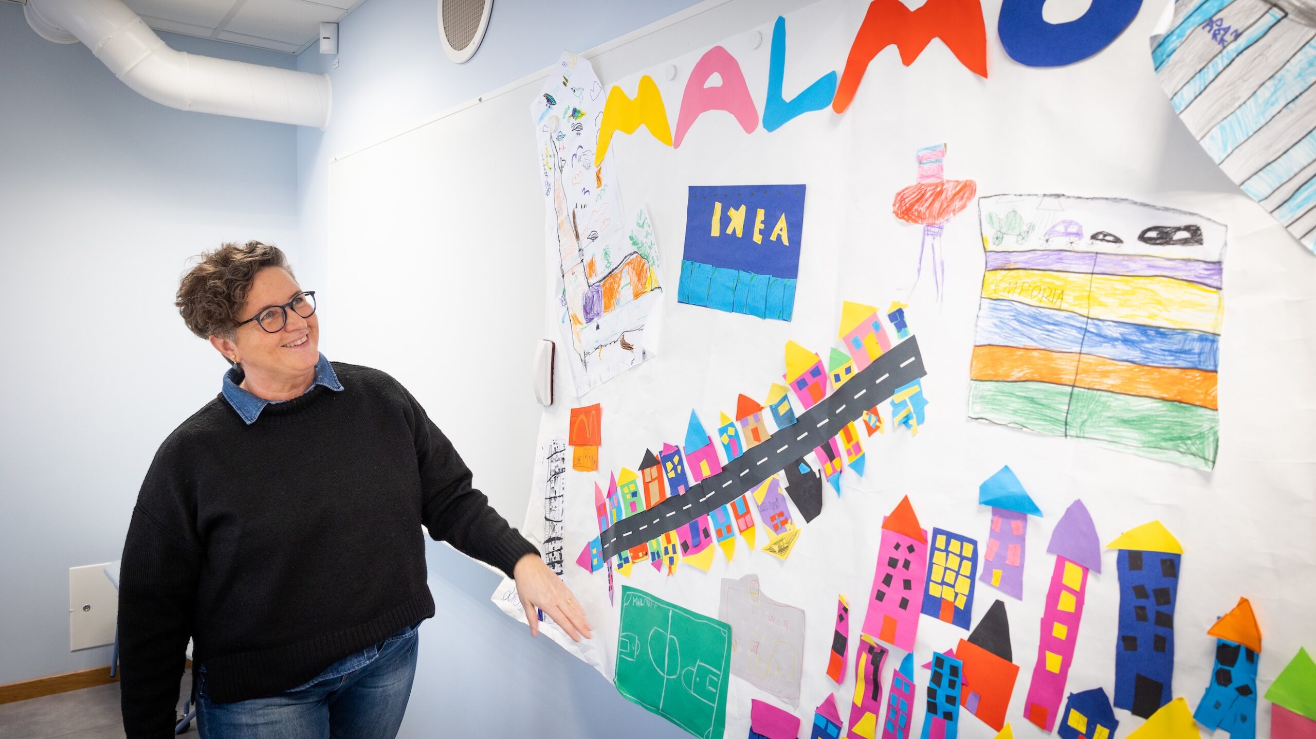 Person står vid en vägg täckt av färgglada barnteckningar med hus, texten Malmö och en IKEA‑logotyp.