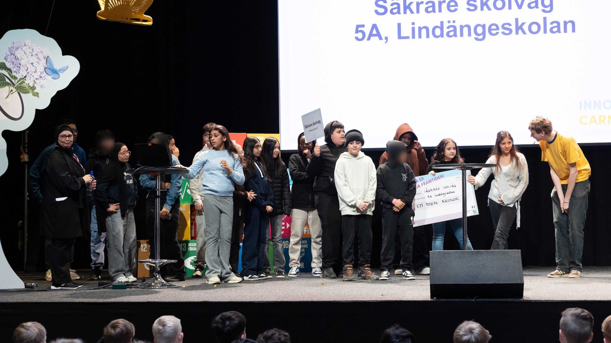 Grupp barn står på en scen framför en stor skärm med texten Säkrare skolväg, 5A, Lindängeskolan.