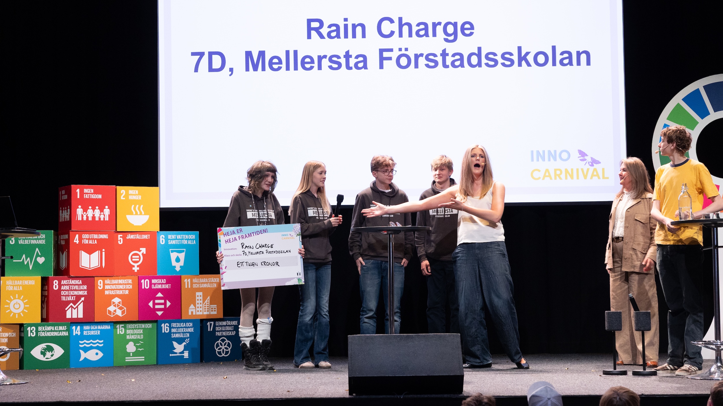 Grupp elever står på en scen framför en stor skärm med texten Rain Charge, 7D, Mellersta Förstadsskolan och InnoCarnival.