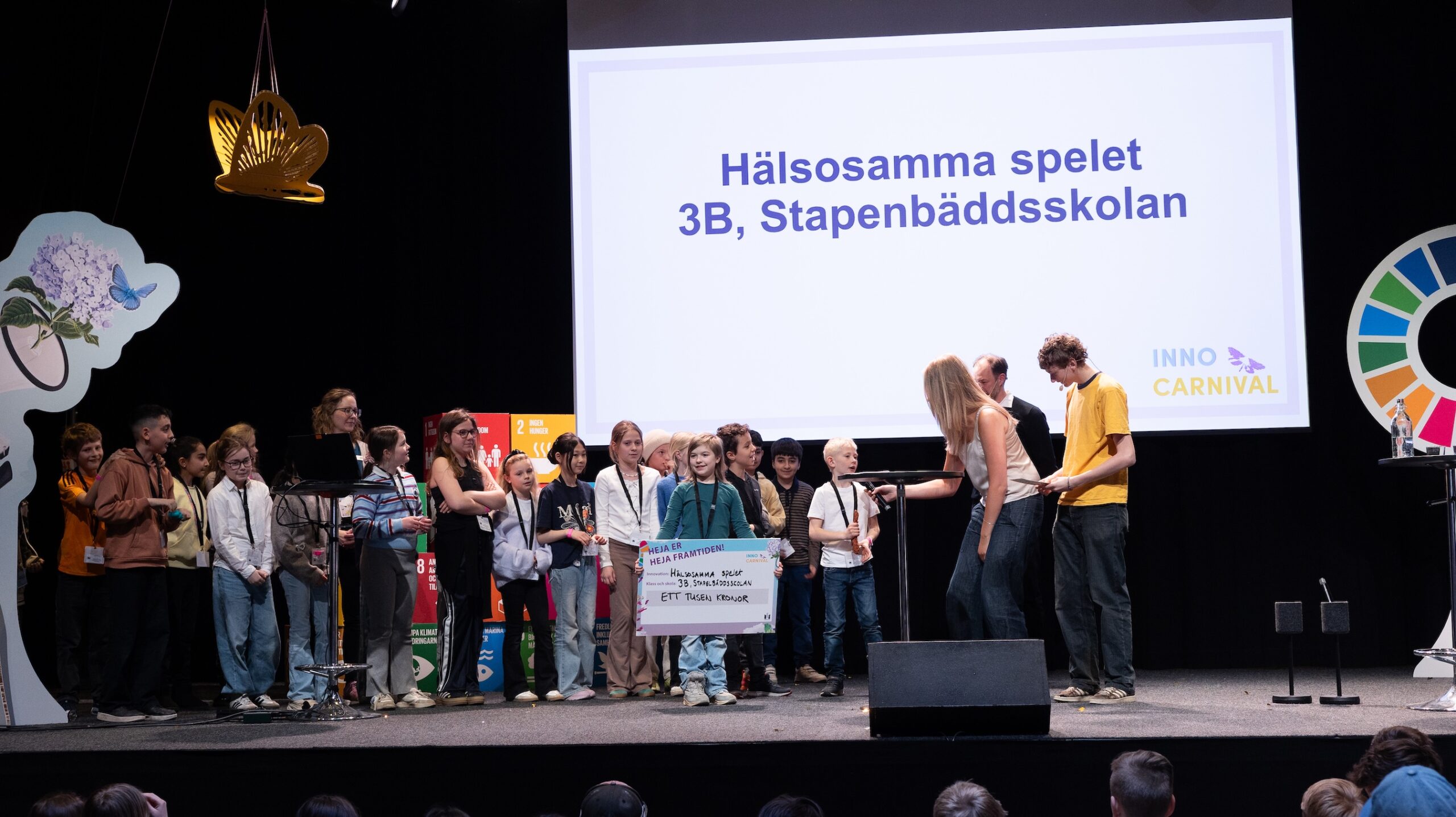 Grupp elever står på en scen framför en stor skärm med texten Hälsosamma spelet, 3B, Stapenbäddsskolan och InnoCarnival.