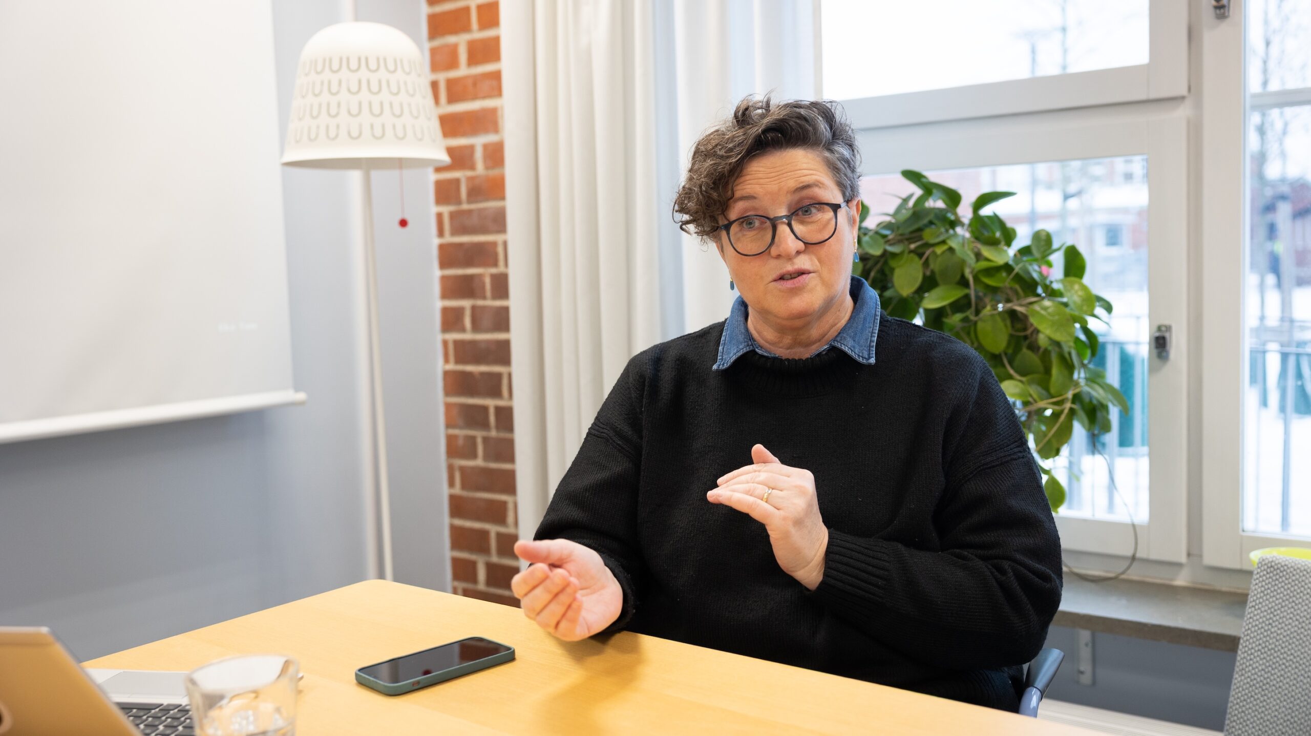 Person sitter vid ett bord och gestikulerar i ett mötesrum med laptop, mobiltelefon och krukväxt vid ett fönster.