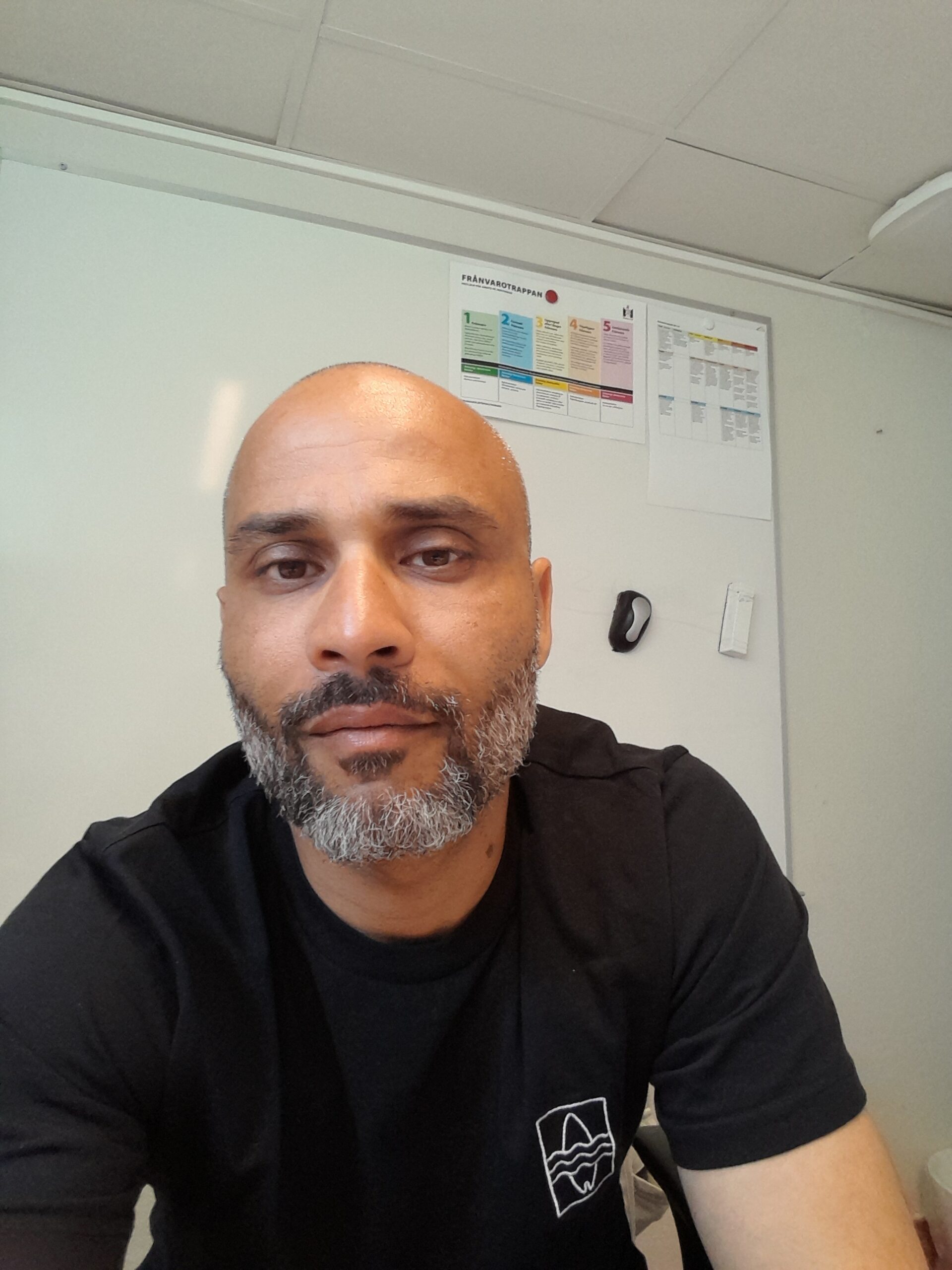 Person med kortklippt hår och skägg sitter i ett rum med vit vägg och anslag bakom.