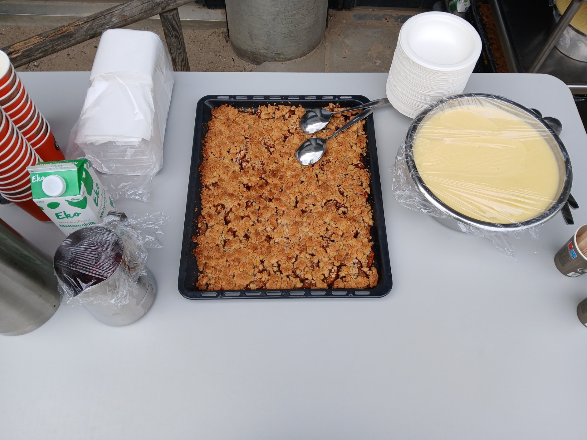 Serveringsbord utomhus med en ugnsform med smulpaj, skål med vaniljsås, papperstallrikar, muggar och mjölk.