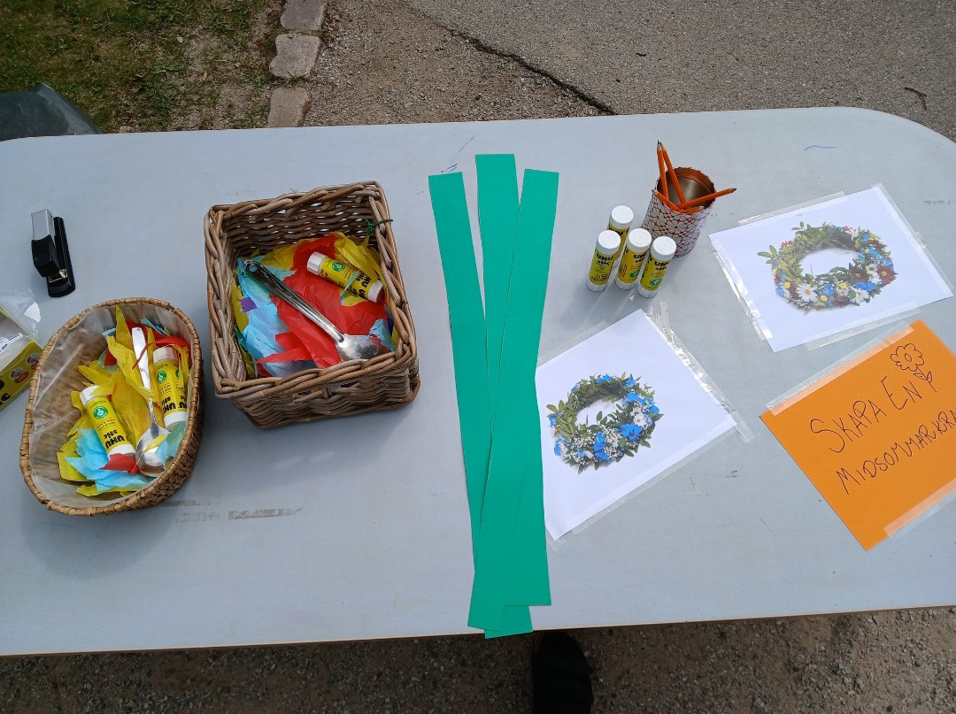 Material för pyssel med pappersremsor, limstift, pennor och bilder av blomkransar, uppställda på ett bord tillsammans med skylt med texten Skapa en midsommarkrans.