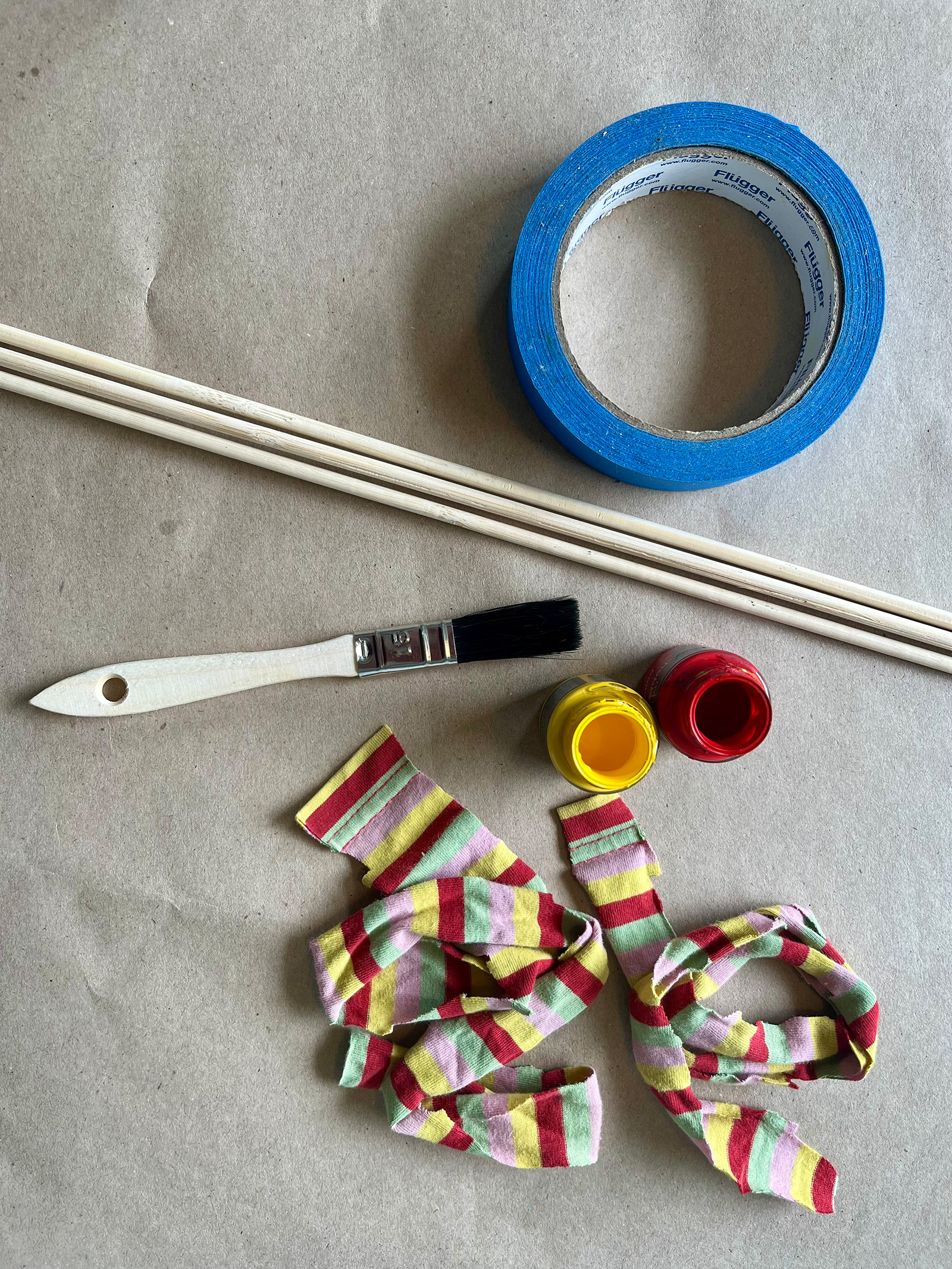 Material för pyssel på bord med träpinnar, pensel, två färgburkar, tygremsor och blå tejprulle.