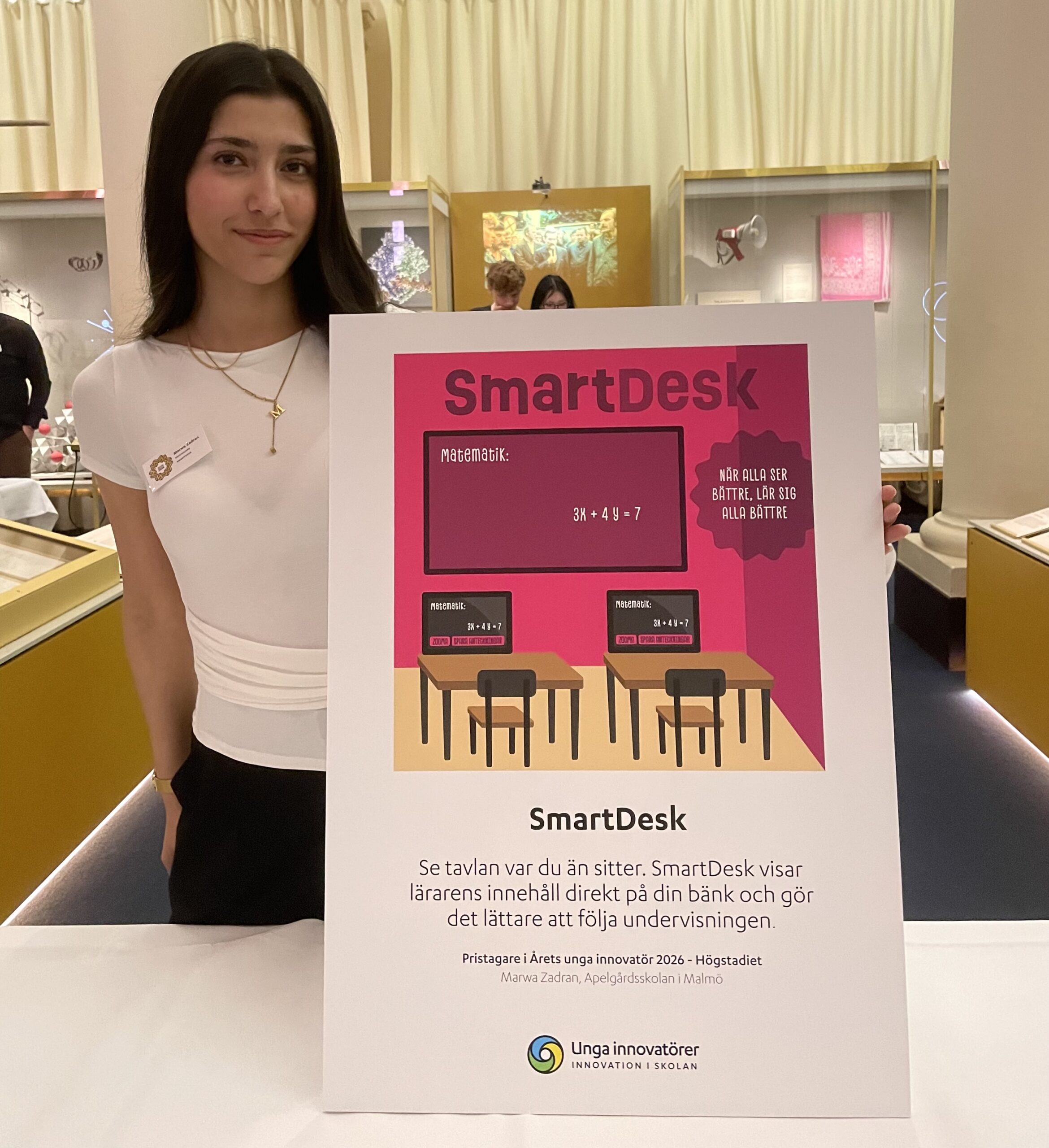 Person står inomhus och håller en affisch med texten SmartDesk om matematik och undervisning, i en utställningsmiljö.