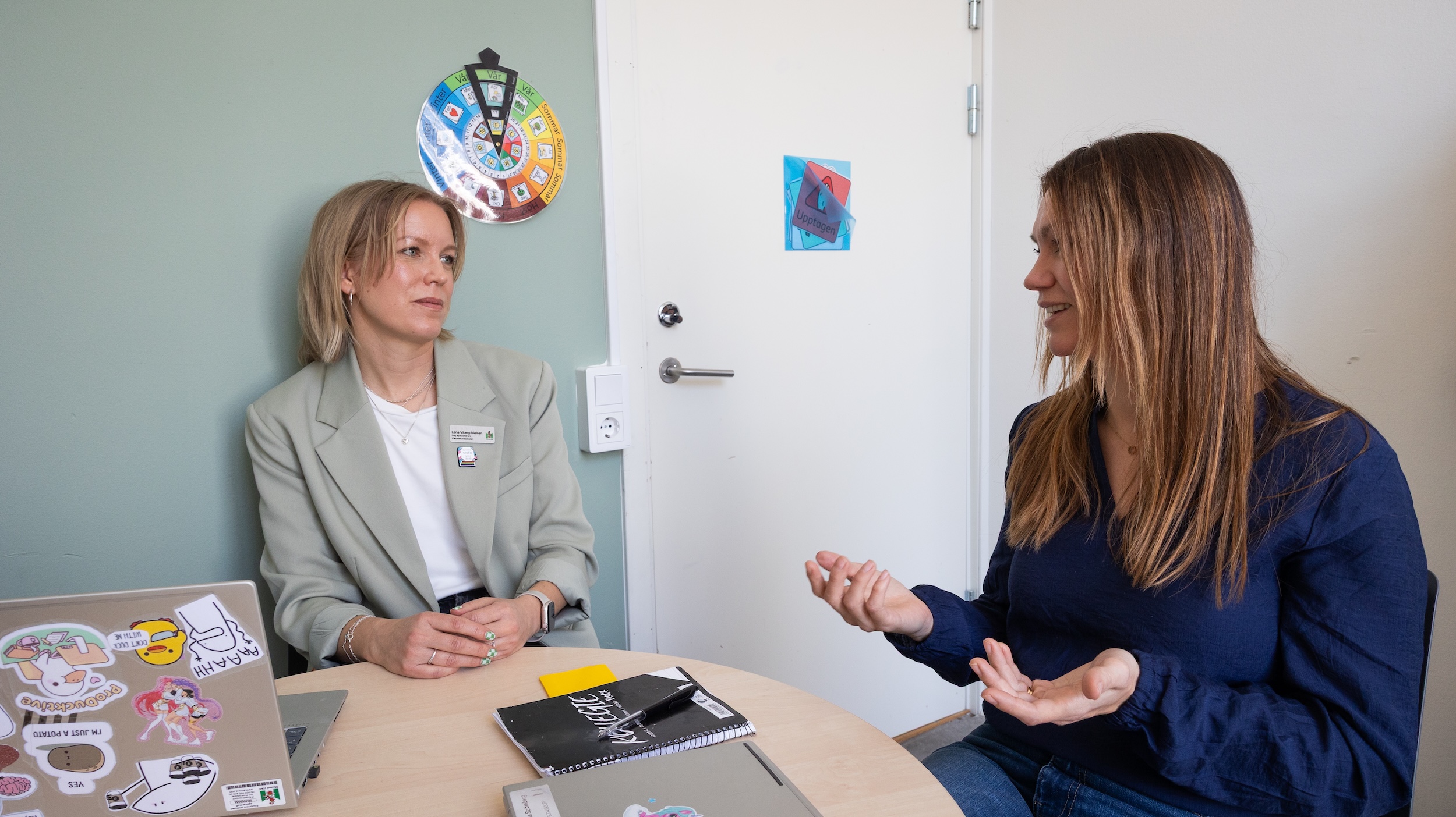 Två personer sitter vid ett runt bord i ett mötesrum och samtalar, med laptop, anteckningsblock och material på bordet.