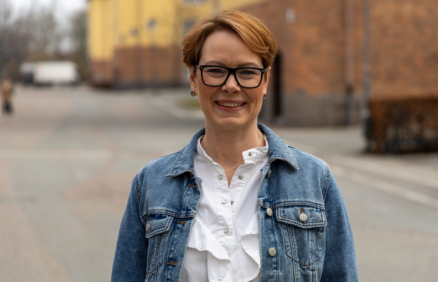 Person står utomhus på en gata framför tegelbyggnader, iklädd jeansjacka och glasögon.