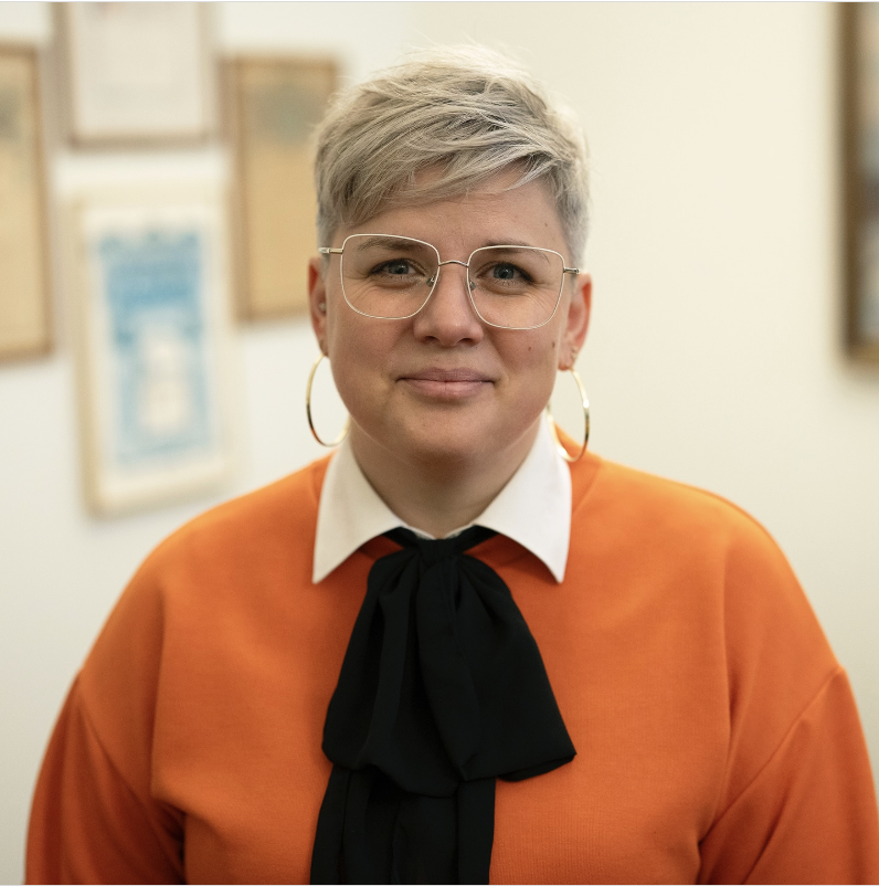 Person med kort hår och glasögon står inomhus framför en ljus bakgrund med tavlor på väggen.