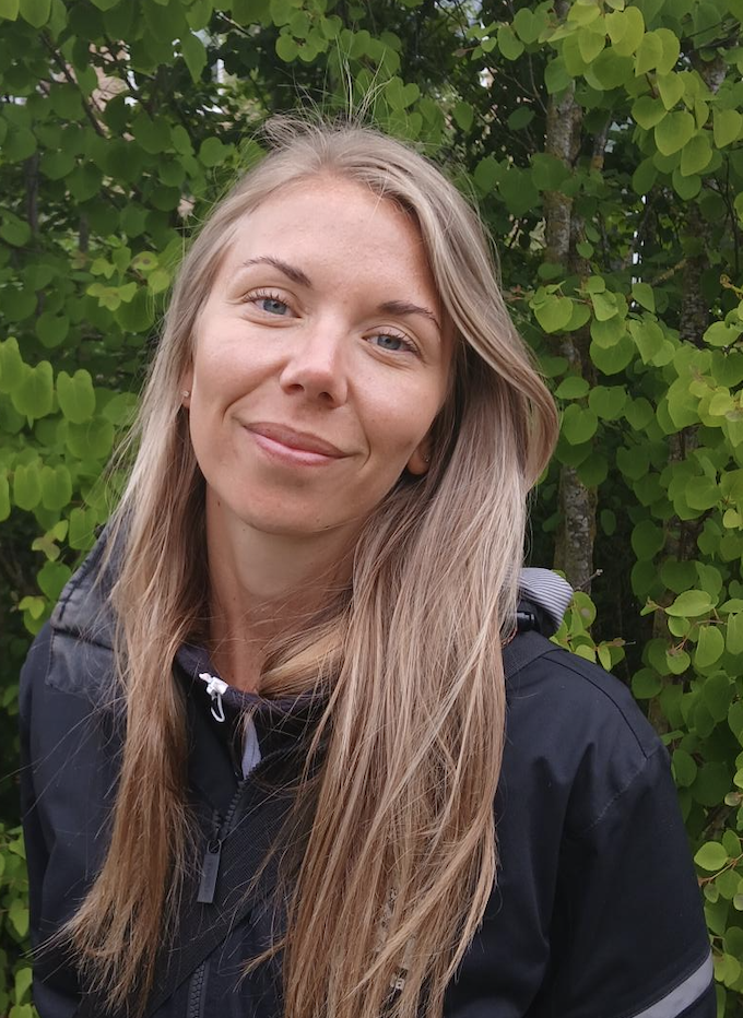 Person står utomhus framför grön buske med löv, iklädd mörk jacka.