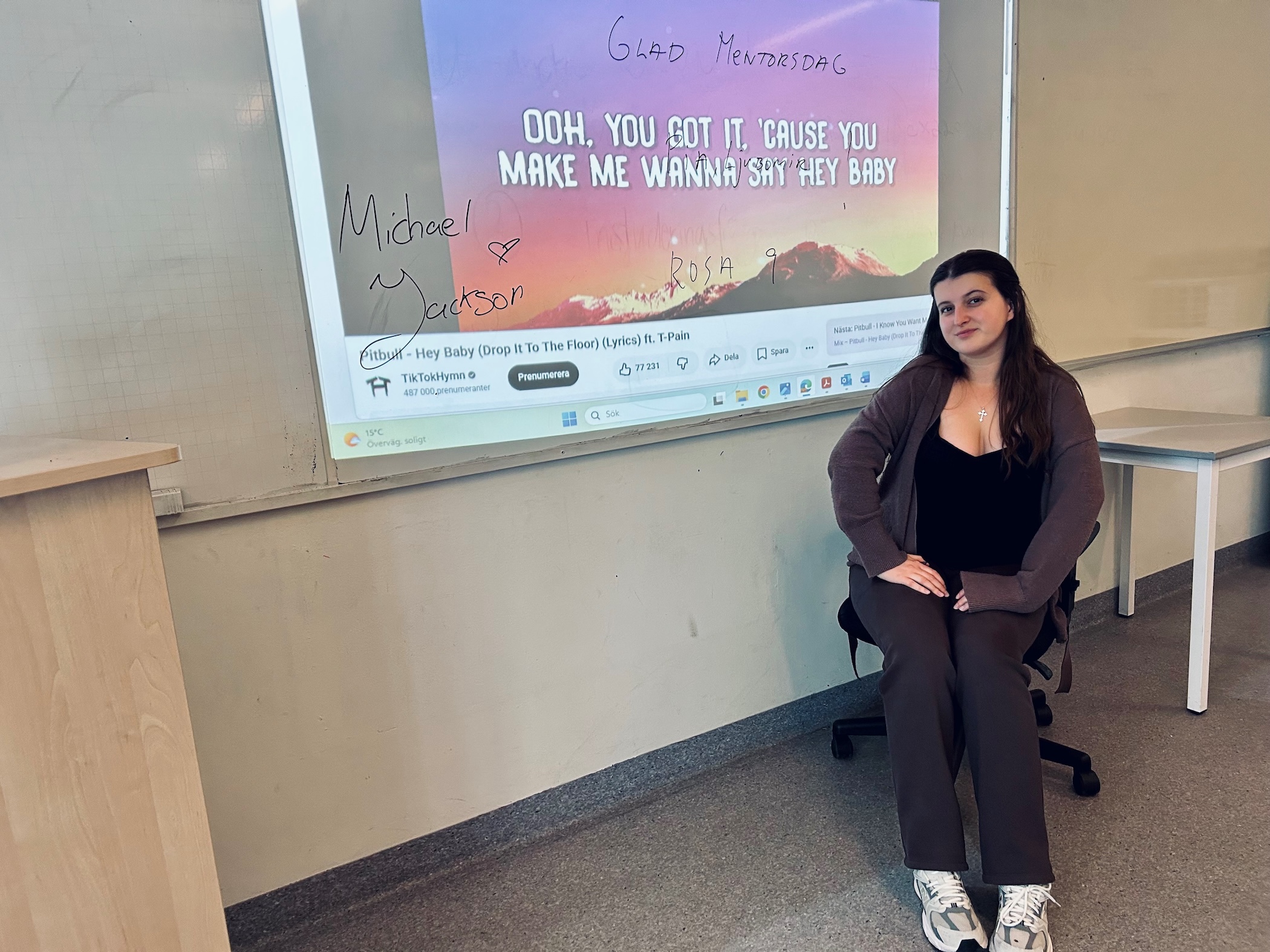 Person sitter på stol i ett klassrum framför en projicerad skärm med låttext och YouTube‑gränssnitt, med handskriven text och namn på whiteboarden runtom.