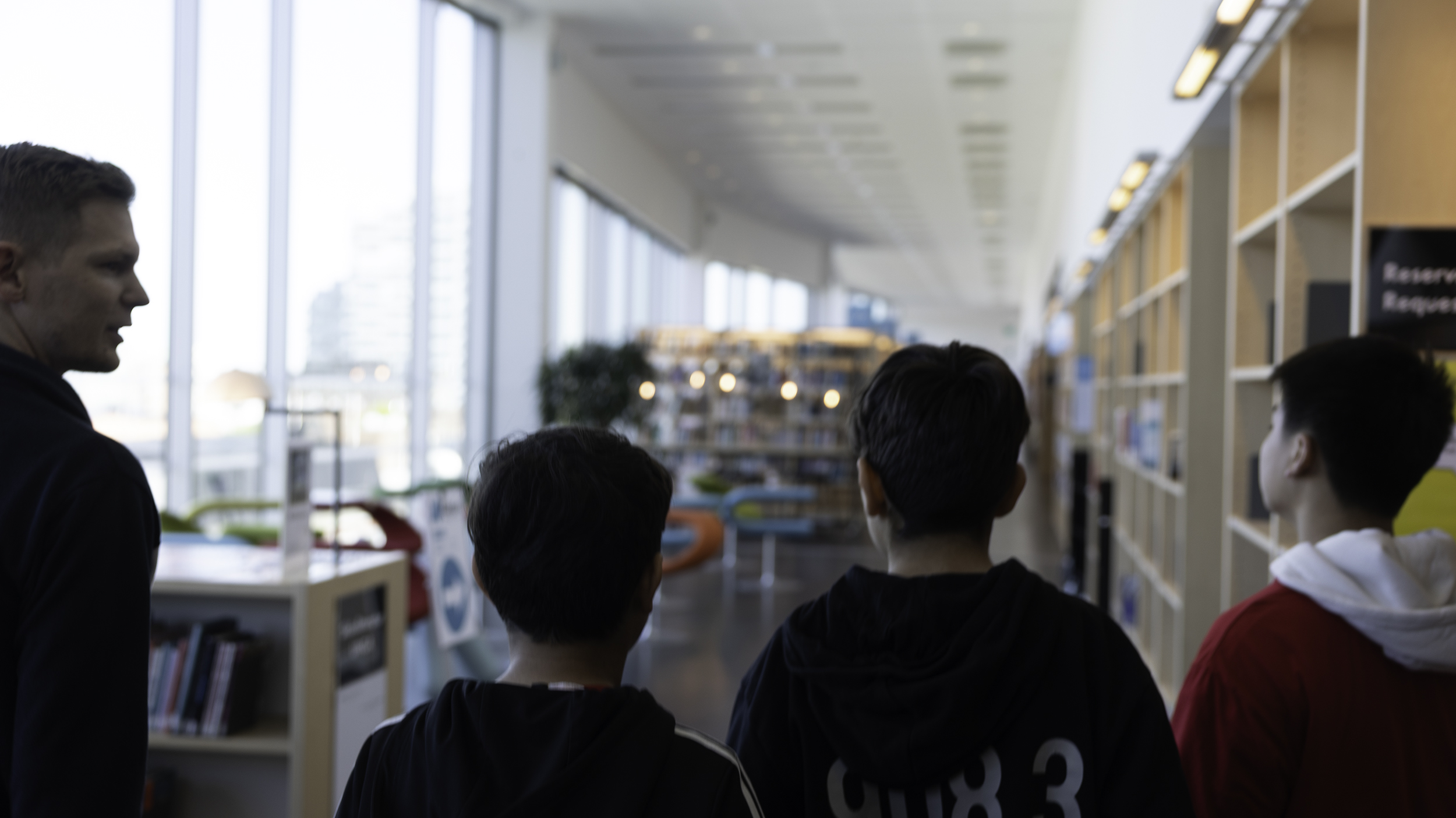 Fyra personer går genom en ljus biblioteksmiljö med bokhyllor, stora fönster och sittplatser i bakgrunden.