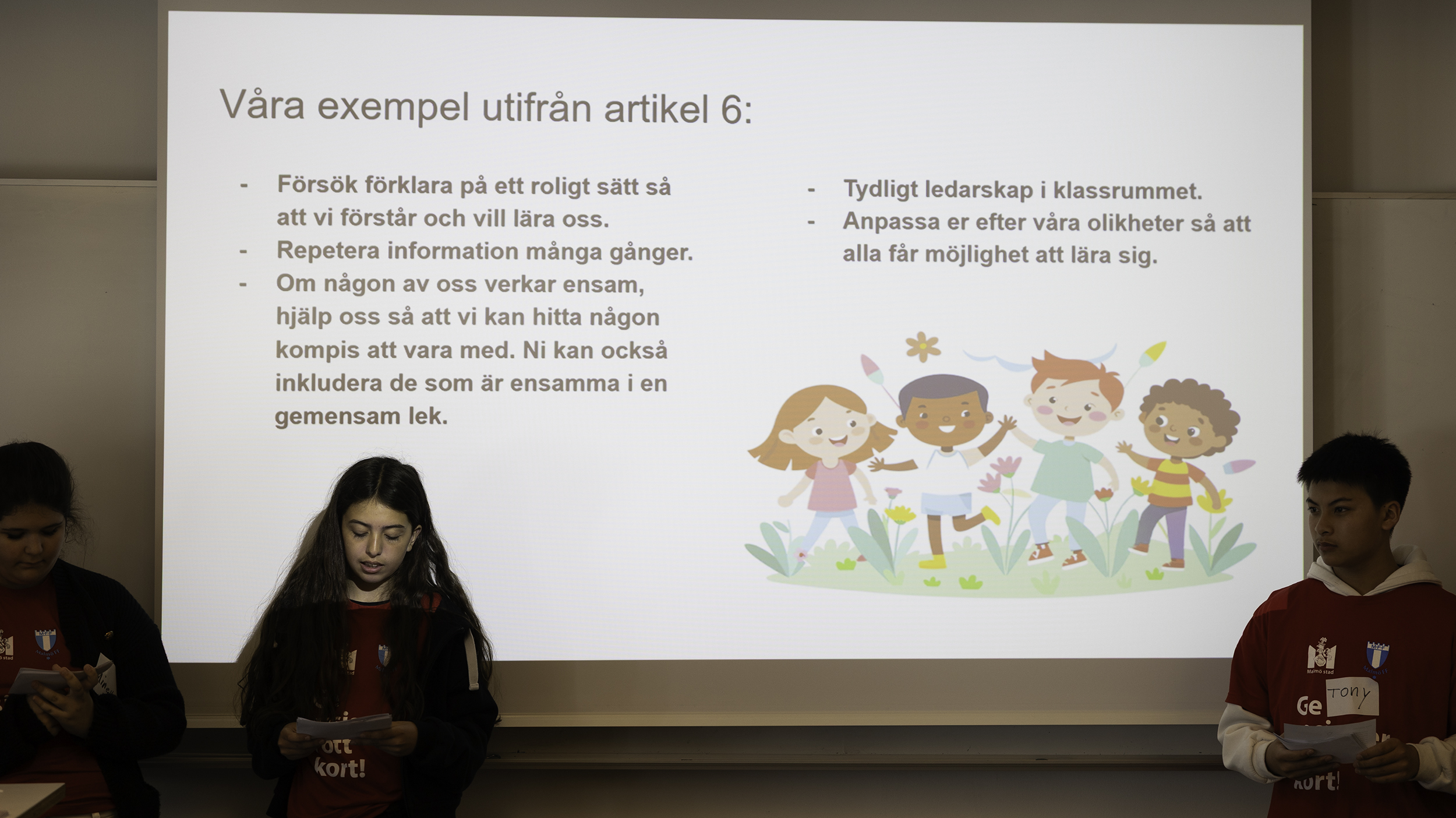 Elever står framför en projicerad presentation med text om artikel 6 och inkludering i klassrummet samt illustration av barn i en blomsteräng.