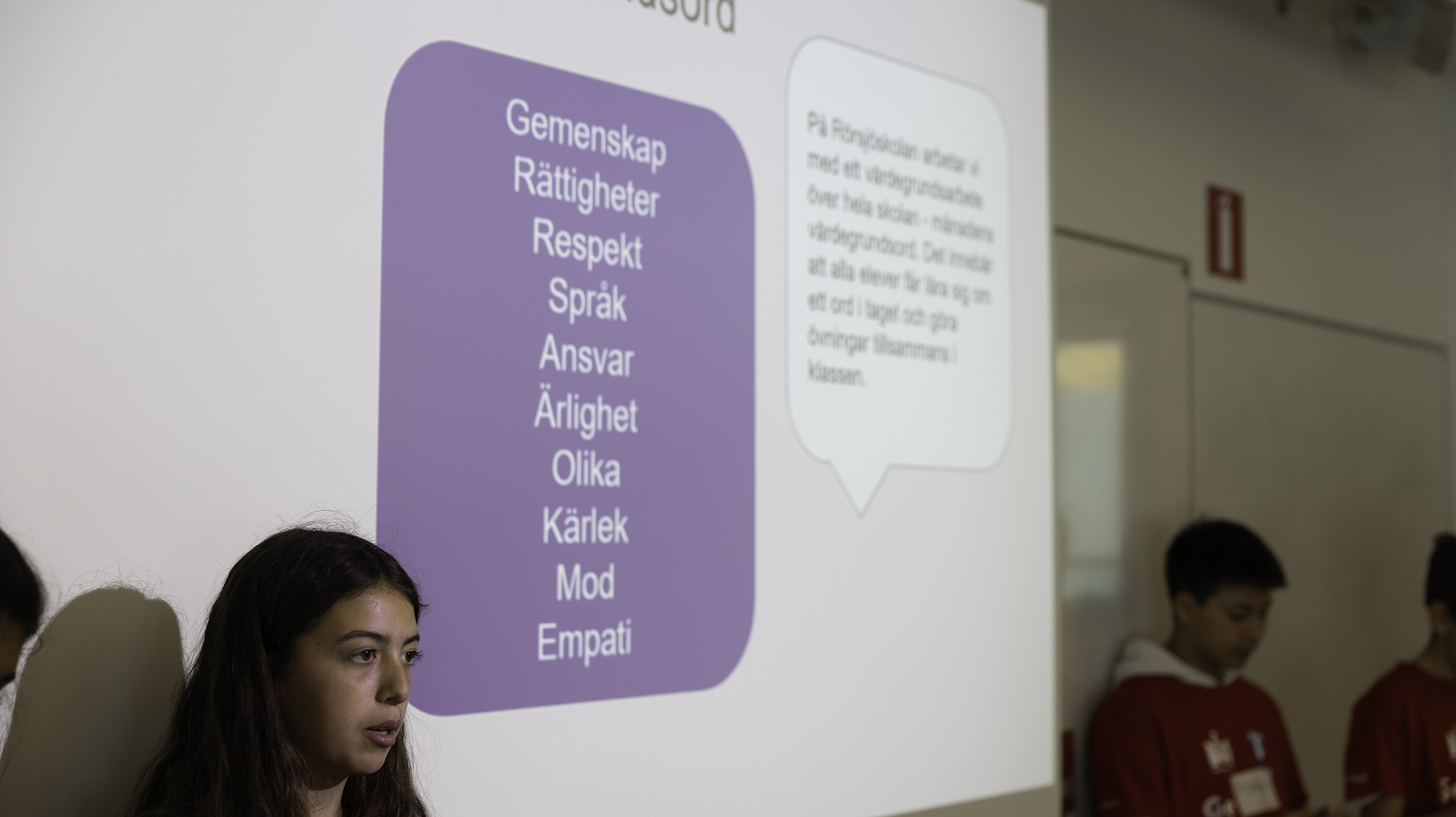 Elev står framför en projicerad presentation med ord som gemenskap, rättigheter, respekt och empati, medan fler elever står bredvid i klassrummet.