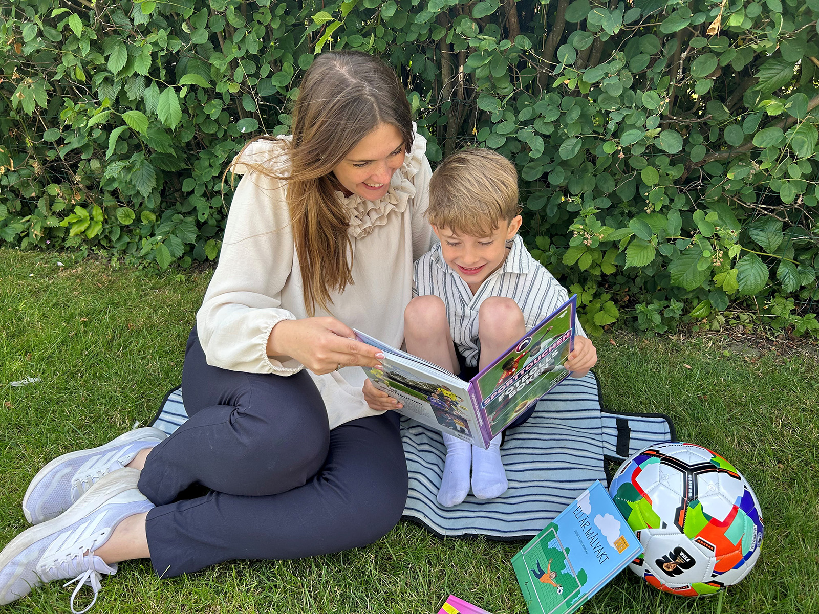 Person och barn sitter på en filt i gräset och läser en bok tillsammans, med en fotboll och fler böcker bredvid samt buskar i bakgrunden.