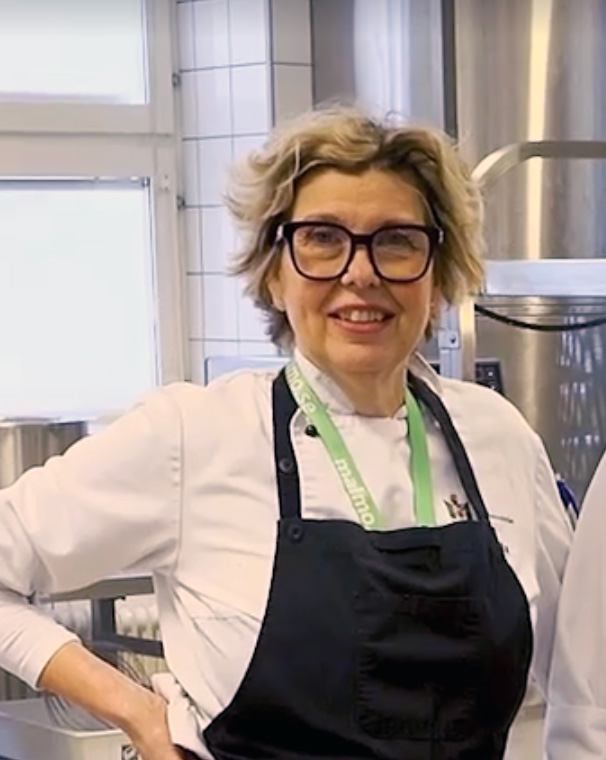 Person med glasögon och förkläde står i ett storkök med rostfri utrustning och kaklade väggar i bakgrunden.