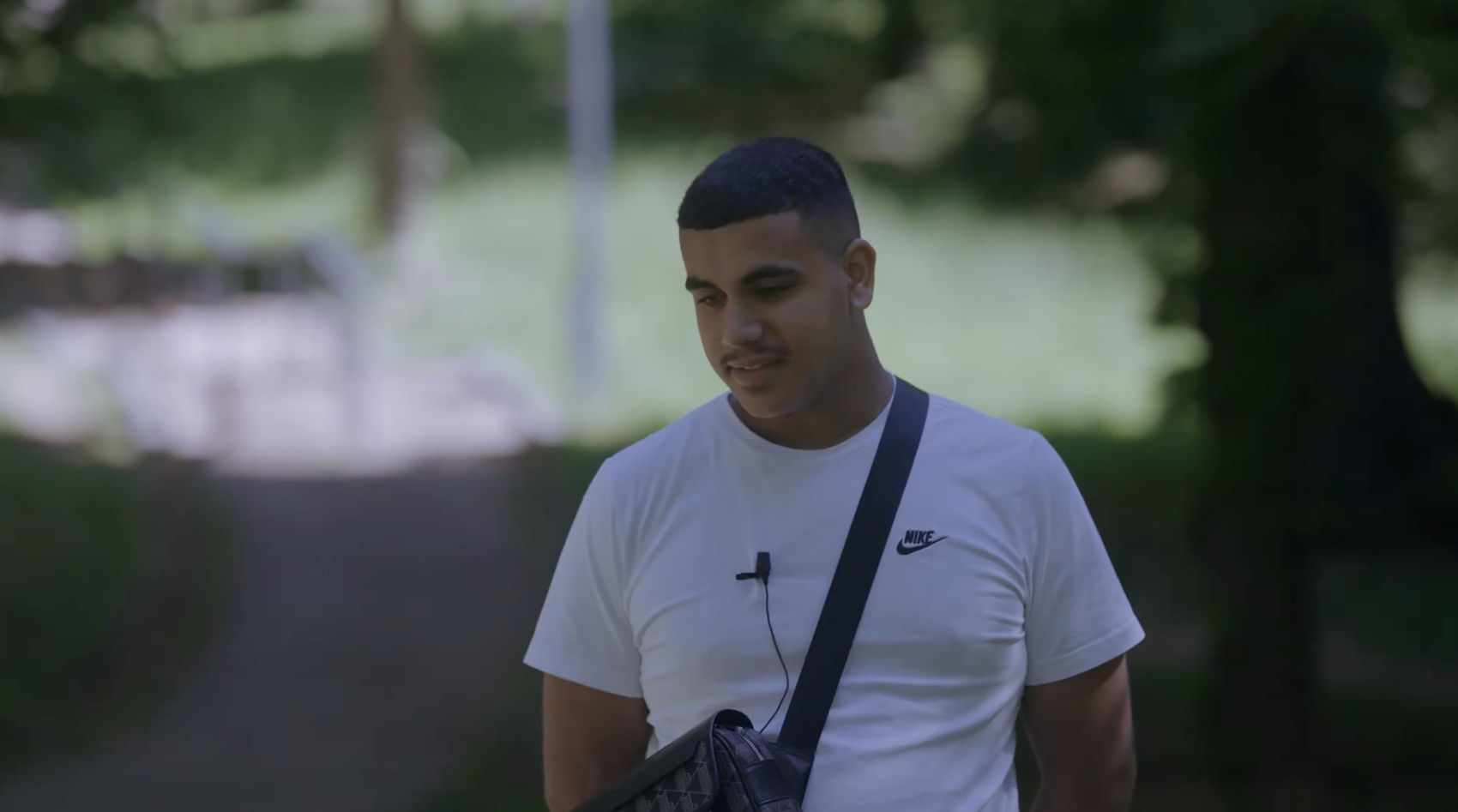Person står utomhus på en gångväg i en parkmiljö, iklädd vit t‑shirt och bär en axelväska med mikrofon fäst på tröjan.
