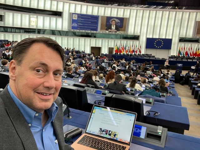 Person sitter vid en laptop i en plenisal med EU‑flagga, nationella flaggor och deltagare samlade framför en stor presentationsskärm i bakgrunden.